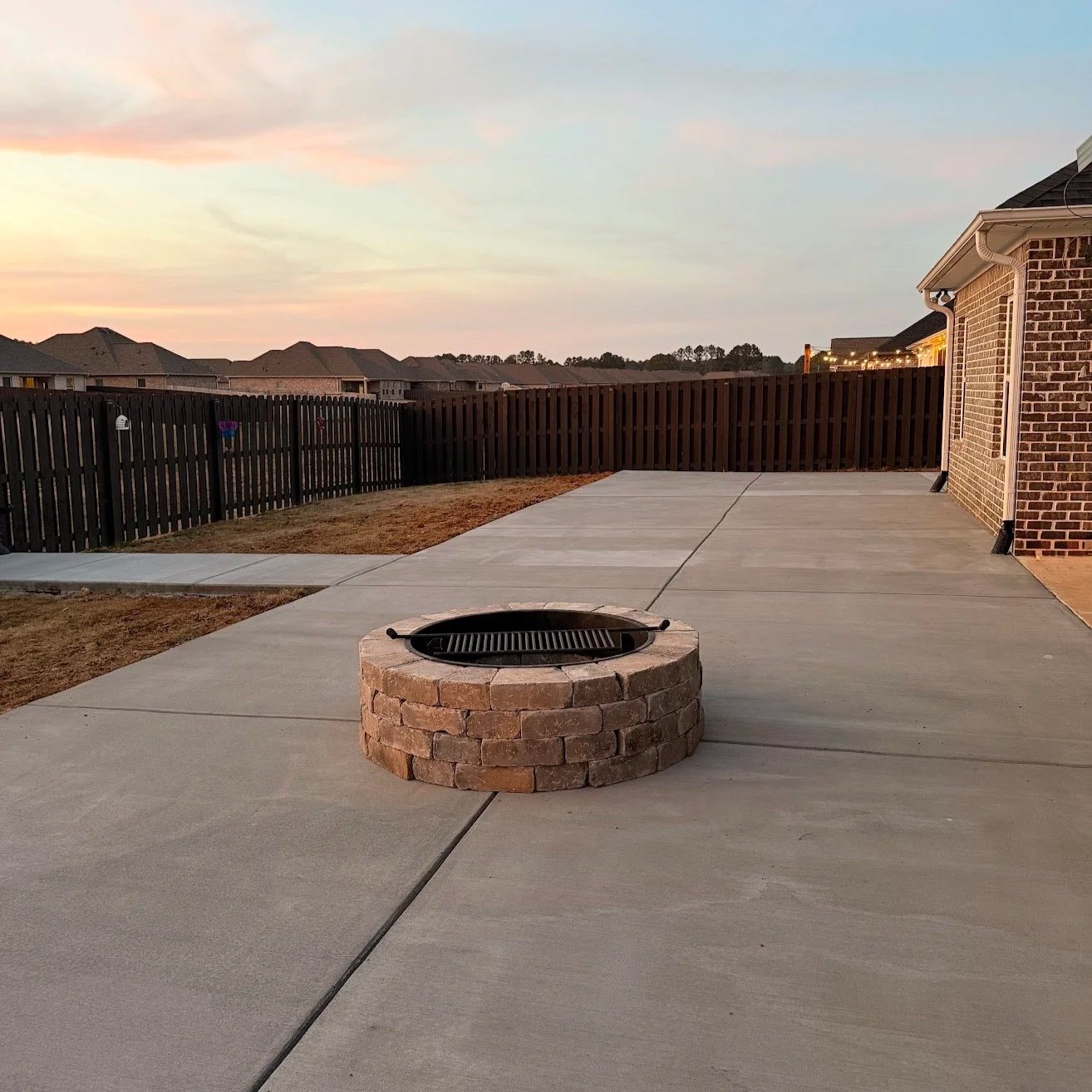 Backyard patio with a brick fire pit, wooden fence, and neighboring rooftops at sunset.