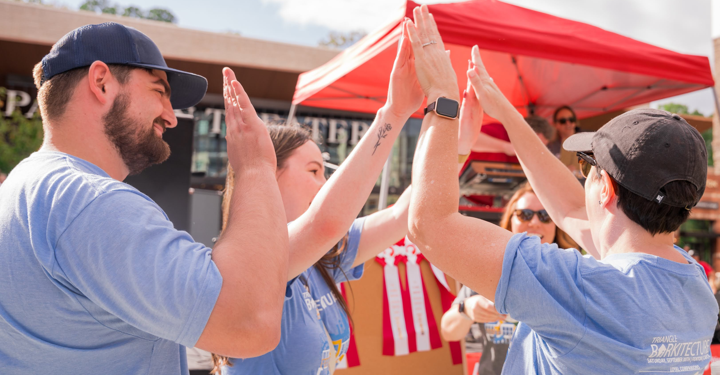 Group of people celebrating outdoors with a high five, standing under a red canopy, wearing matching blue T-shirts.