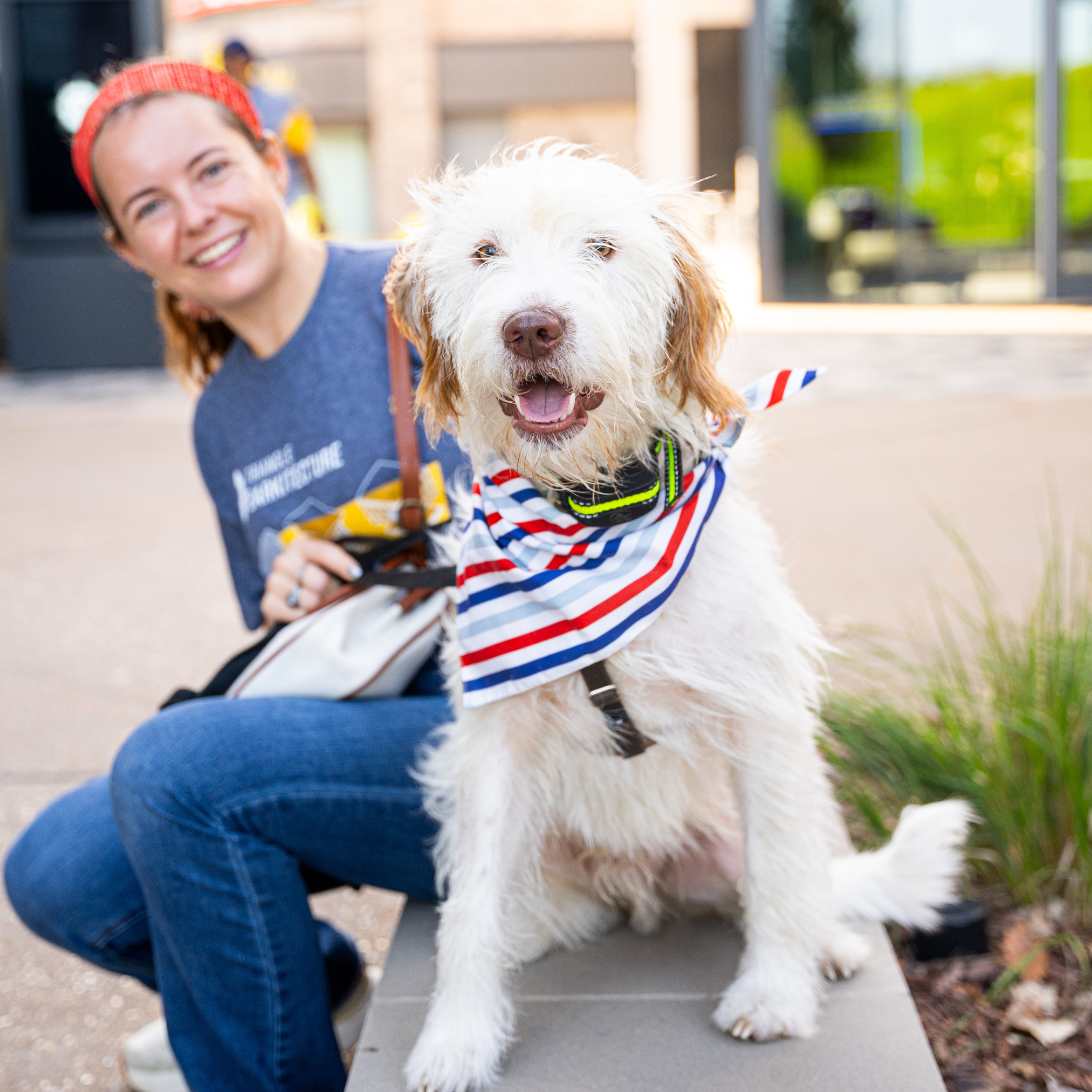 A woman with a red headband sitting beside a white dog wearing a patriotic bandana, outside a modern building.