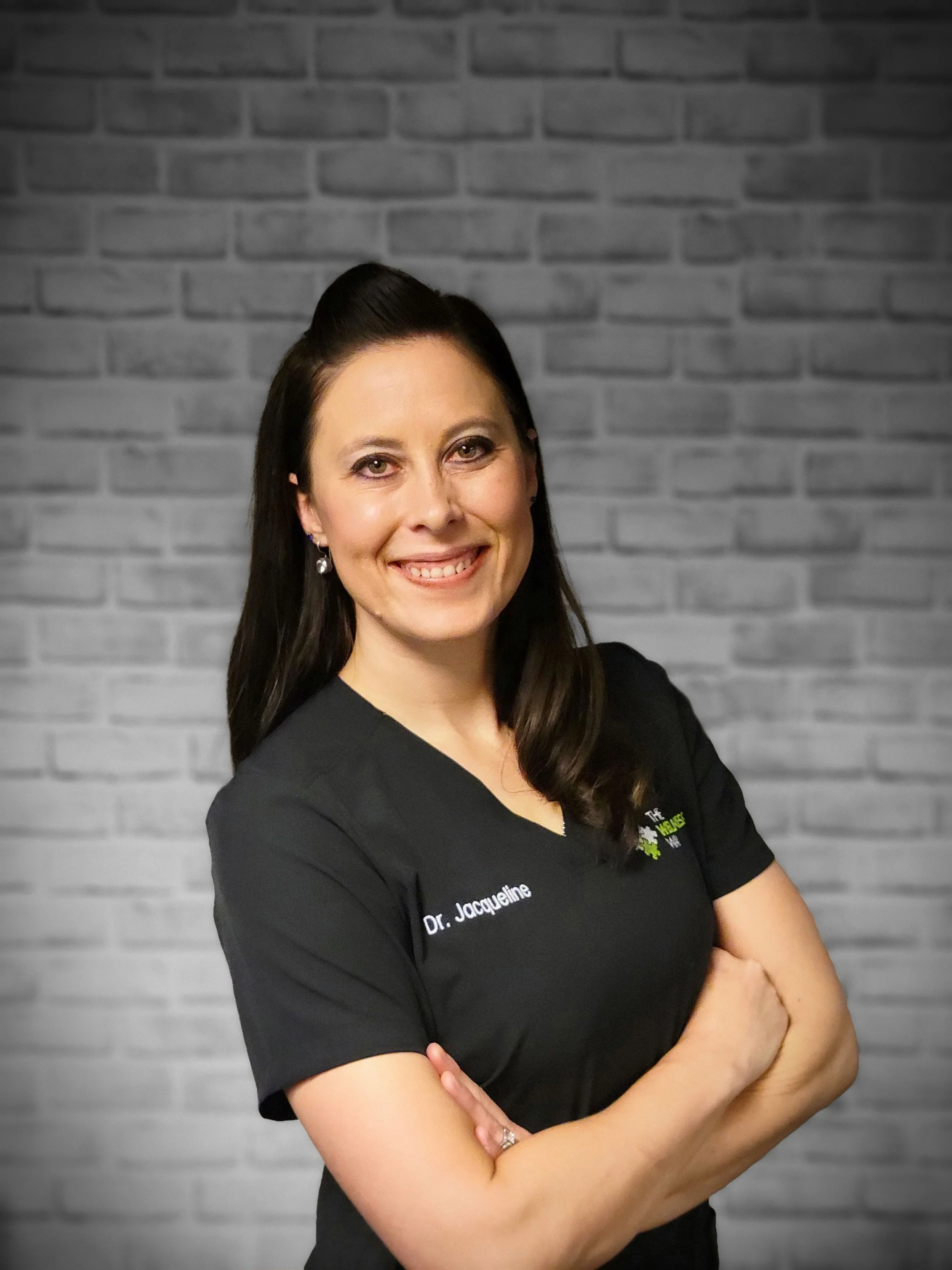 Woman in black medical uniform smiling, standing against a brick wall background.