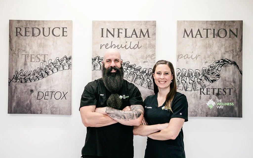 Two people standing in front of three posters with spine illustrations, text includes "REDUCE TEST DETOX," "INFLAM rebuild," and "MATION pair RETEST," promoting The Wellness Way.