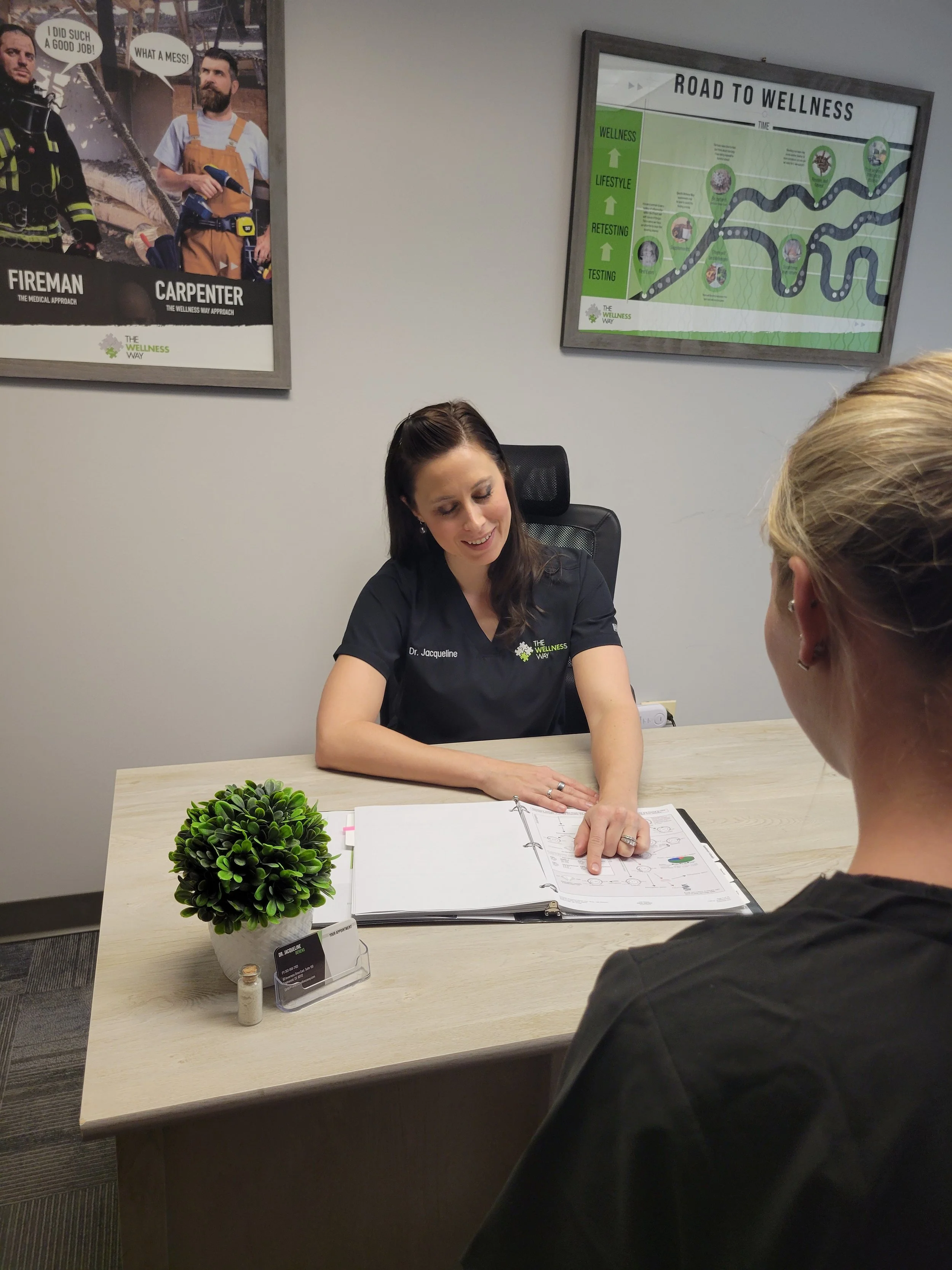 Consultation in a wellness office, a woman showing a binder with documents to another person across a desk, with wellness-themed posters on the wall.