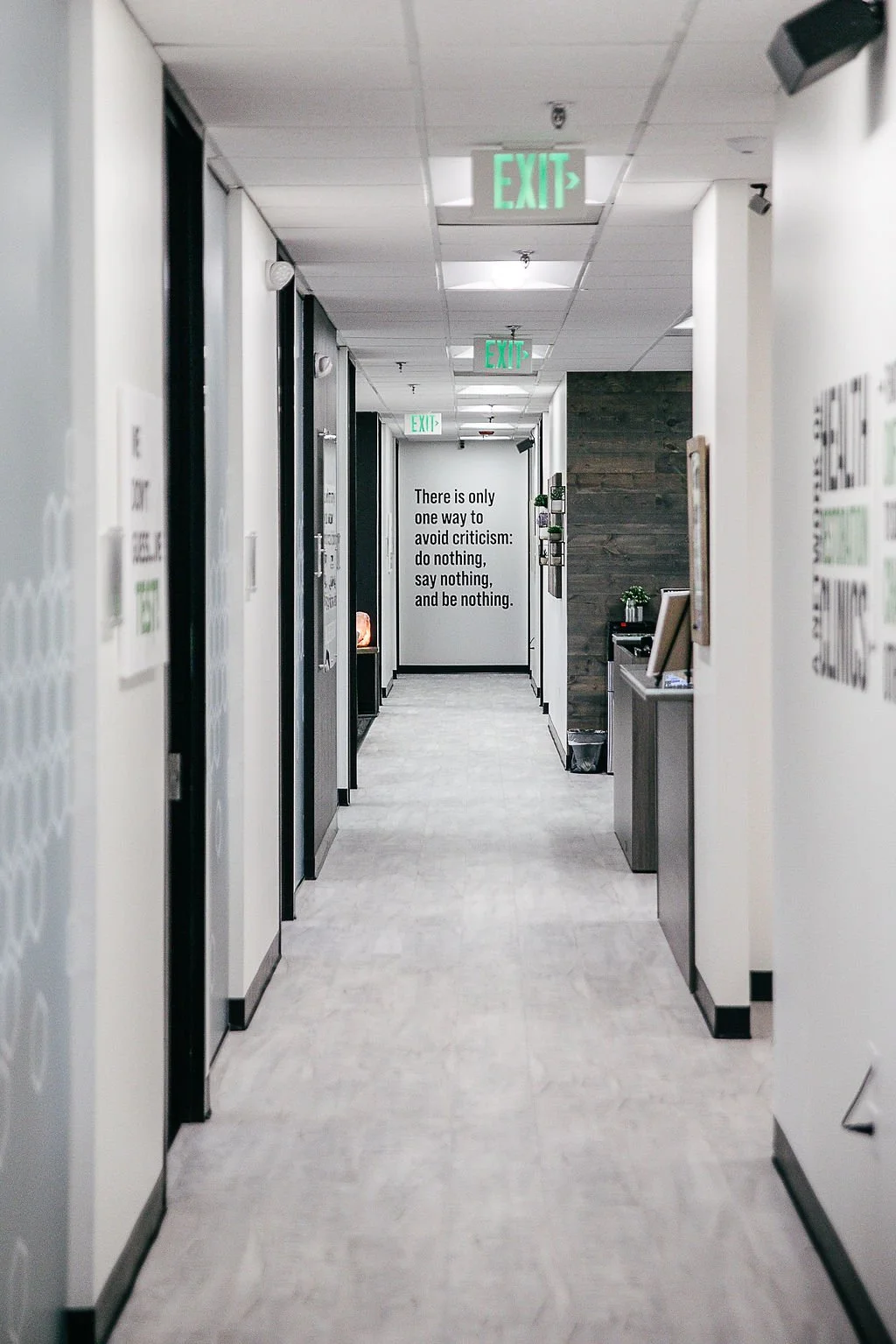 Office hallway with several doors and an exit sign. A motivational quote on the wall reads: 'There is only one way to avoid criticism: do nothing, say nothing, and be nothing.'