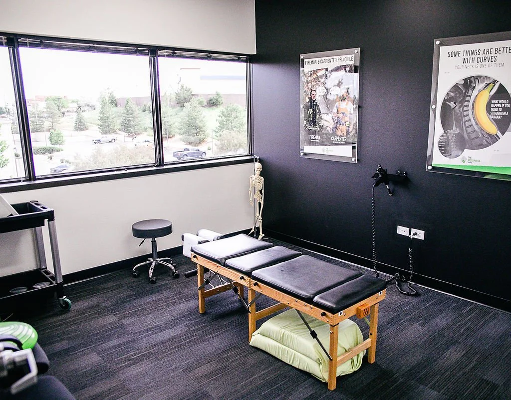Chiropractor office interior with adjustment table, stool, skeleton model, posters on black wall, and large windows.