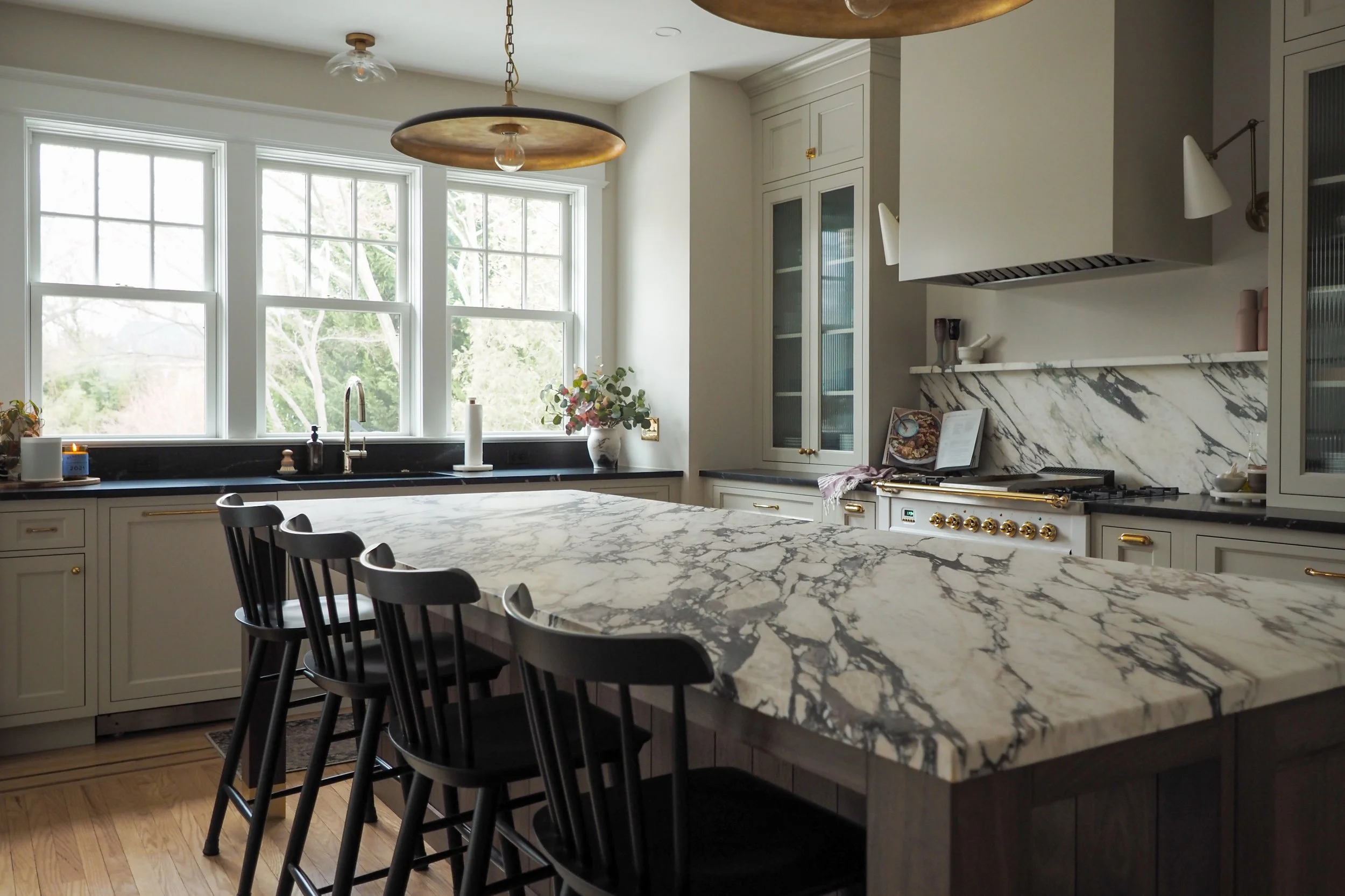 Bright kitchen with marble island, black Windsor stools, brass pendant, and professional range — Studied Milieux interior design