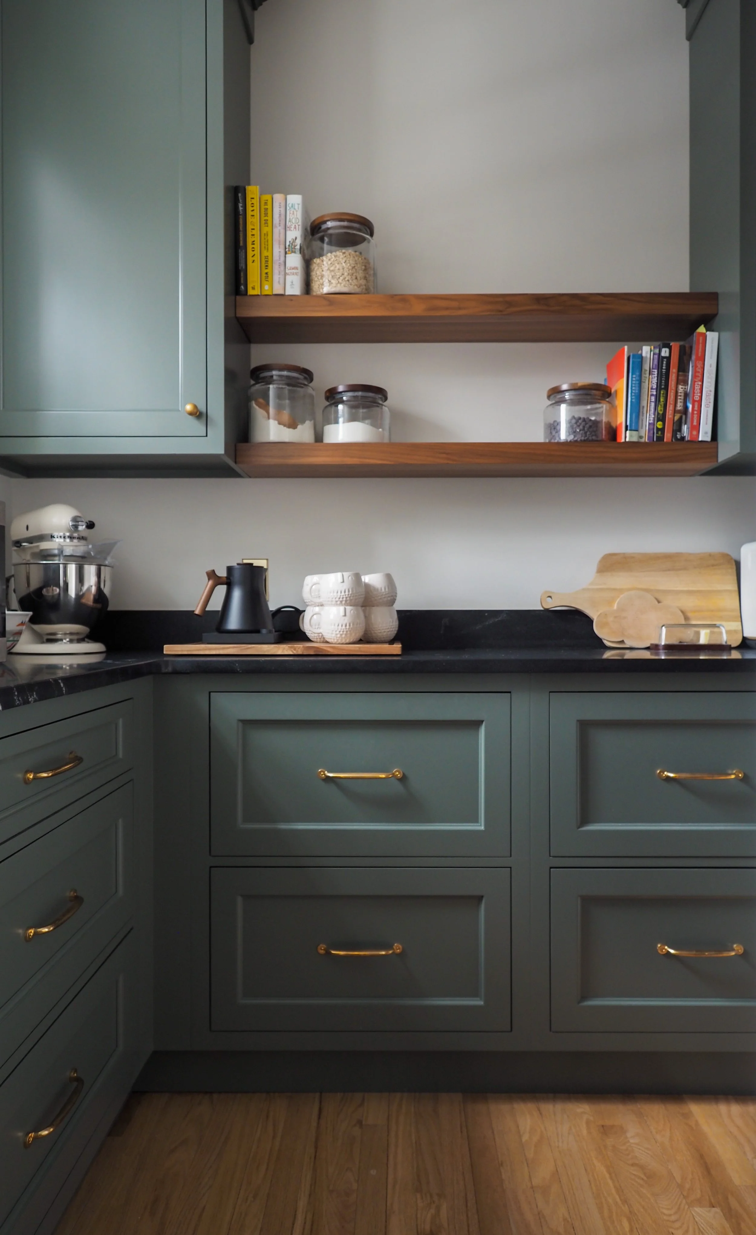 Deep green kitchen cabinetry with open wood shelving and brass hardware — Studied Milieux interior design, South Jersey