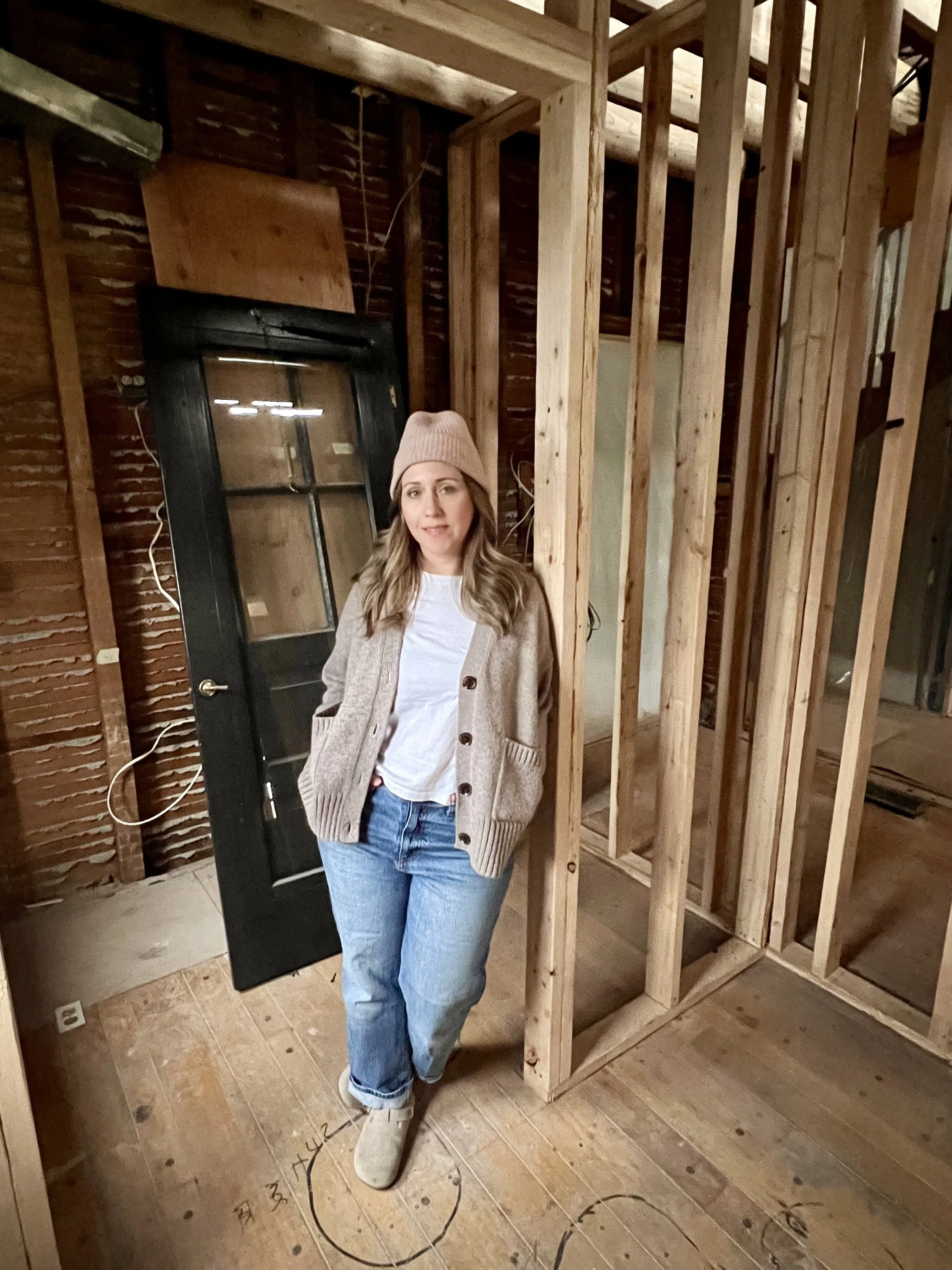 A woman standing inside a partially under construction room with exposed wooden framing and a black door leaning against the wall.