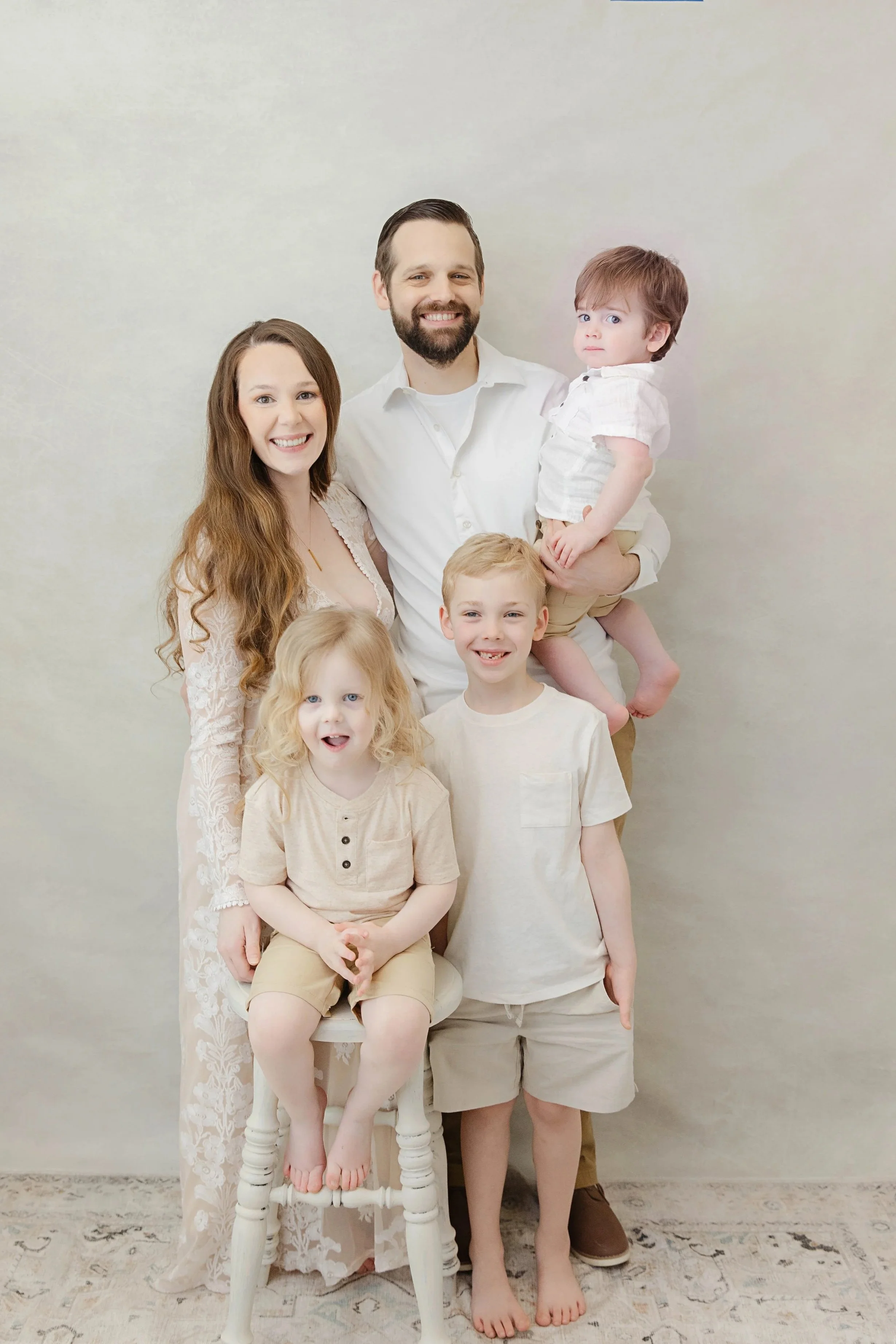 Family portrait of six people, including a woman, man, and four children, in light-colored clothing, standing against a plain light background.