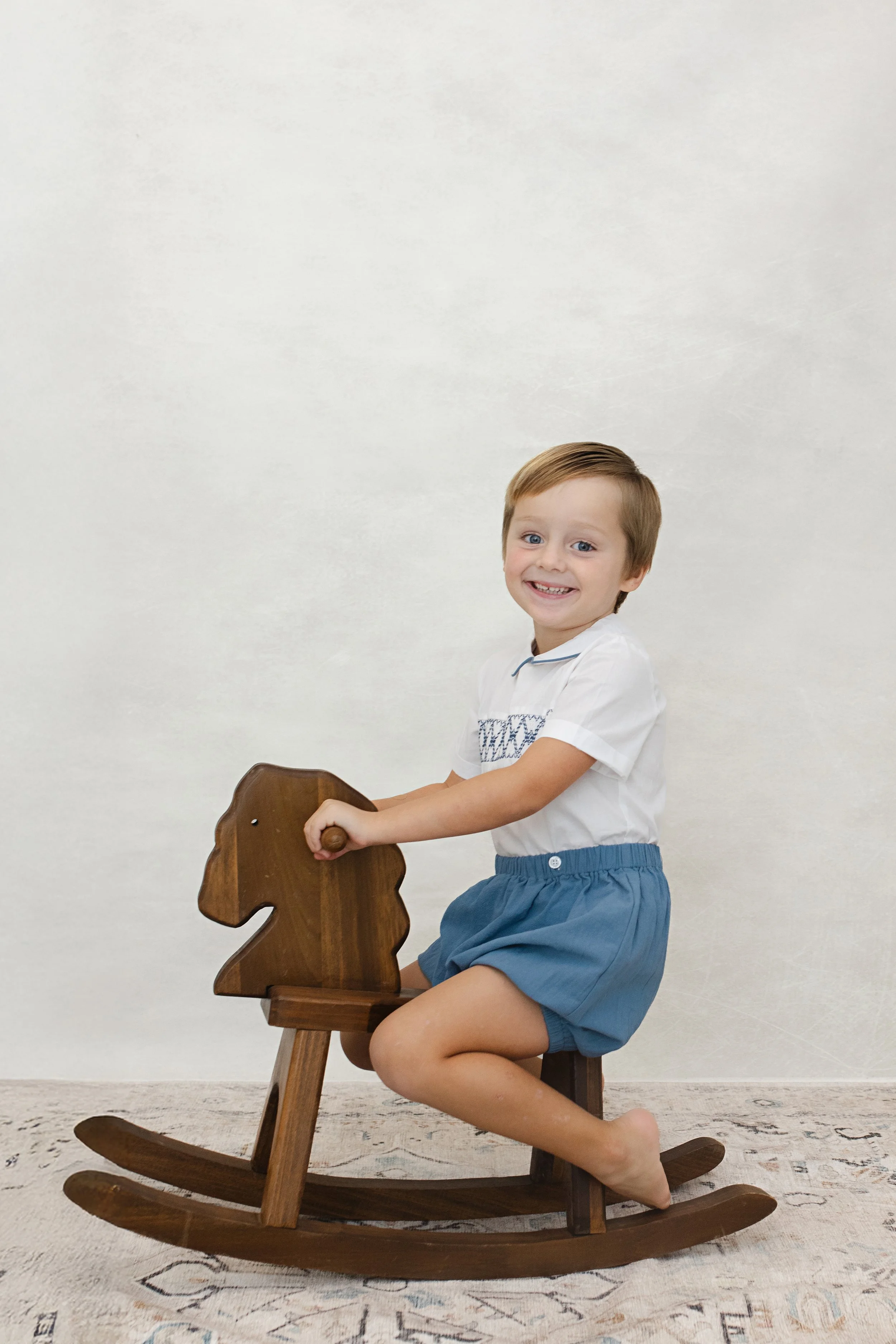 A young boy with brown hair, smiling, riding a wooden rocking horse indoors on a patterned rug against a plain white wall.