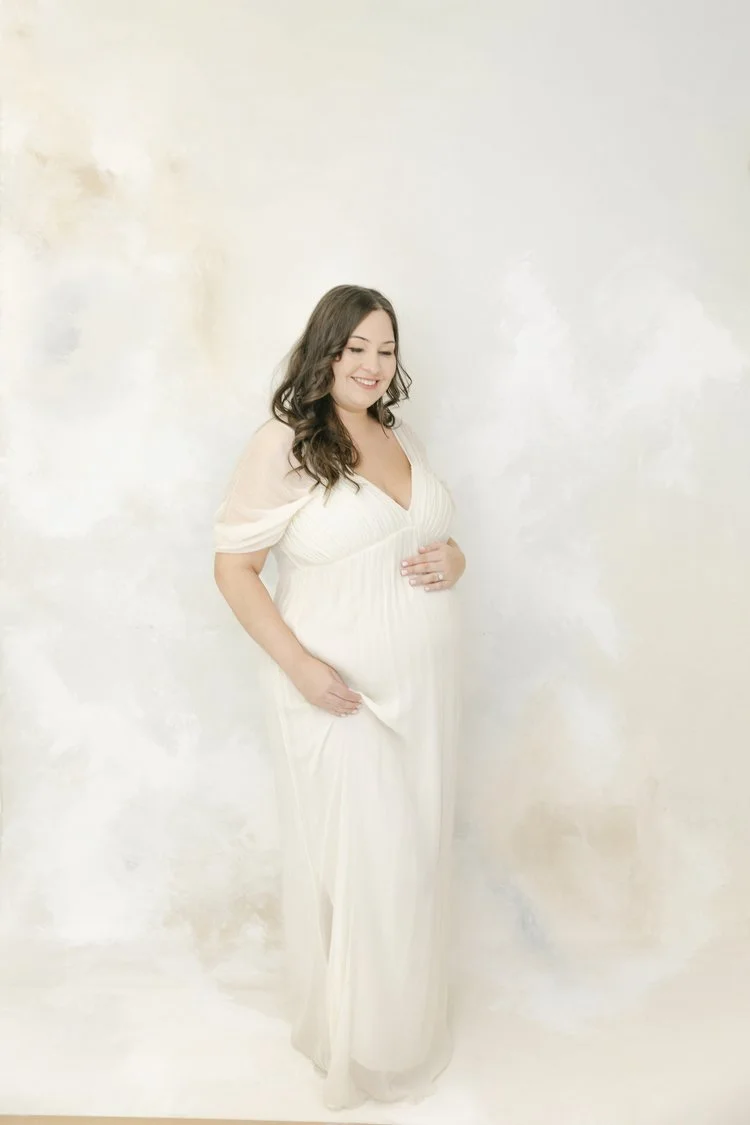 A pregnant woman with long dark hair smiling, dressed in a flowing white maternity dress, standing against a plain light-colored wall.
