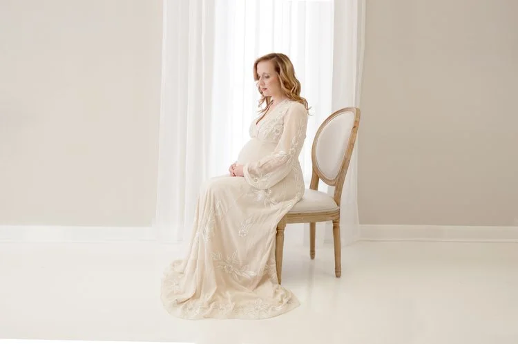 A pregnant woman sitting on a wooden chair in a bright room with white curtains and walls, wearing a long, white lace dress.