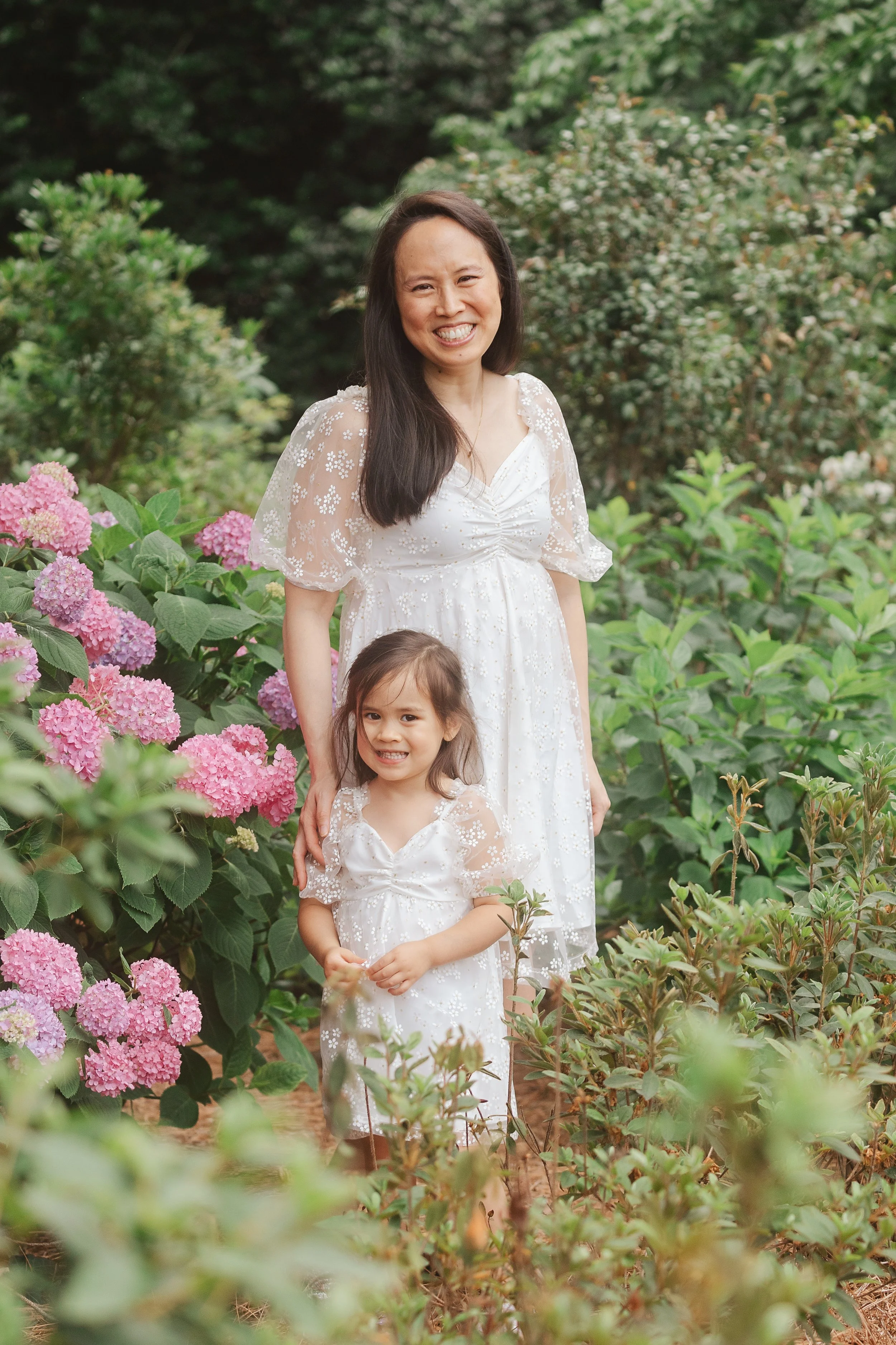 Woman and young girl smiling and standing in a garden with pink hydrangea flowers, both wearing white dresses with floral patterns.