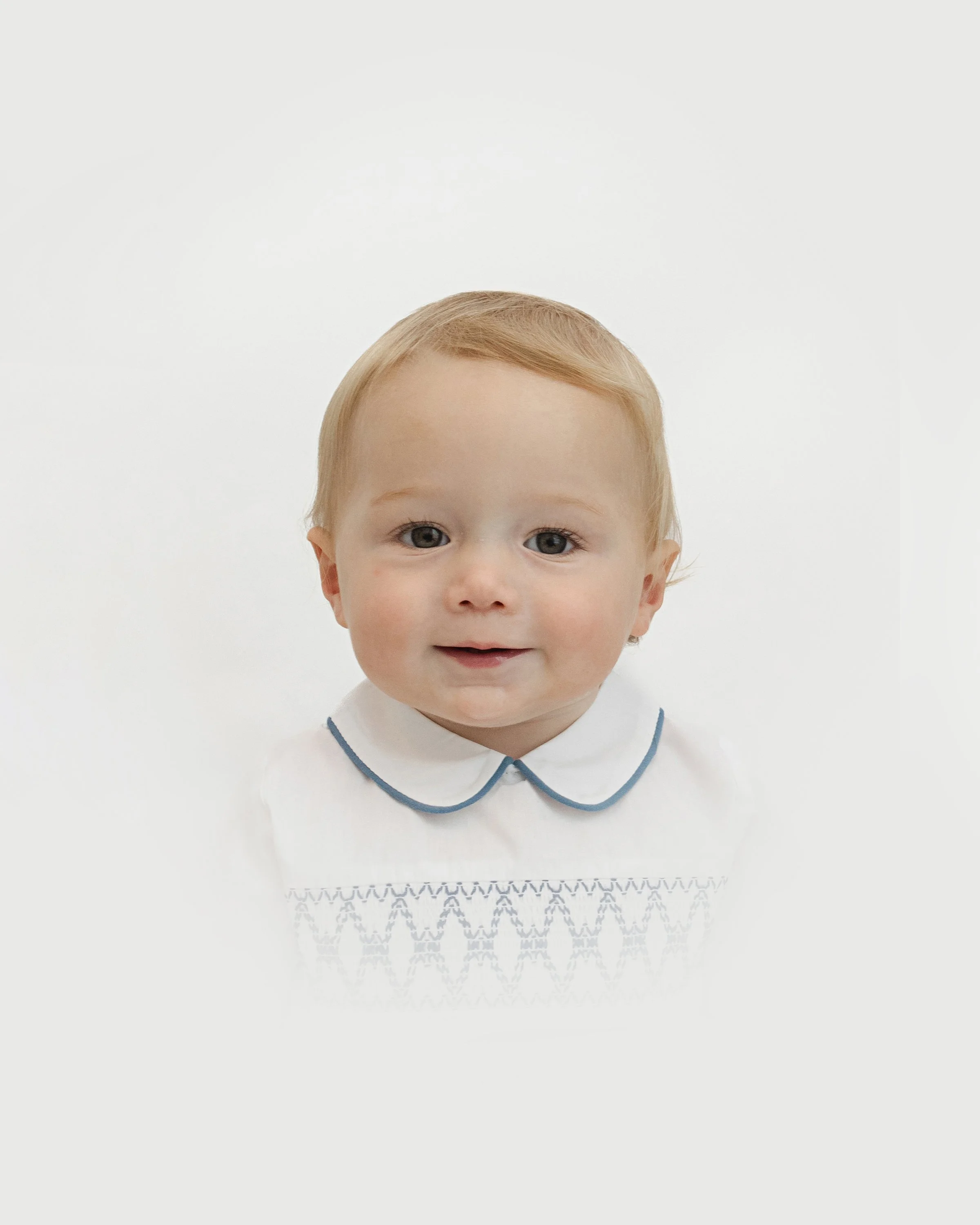 Young child with blond hair and a white shirt with a Peter Pan collar, smiling against a plain white background.