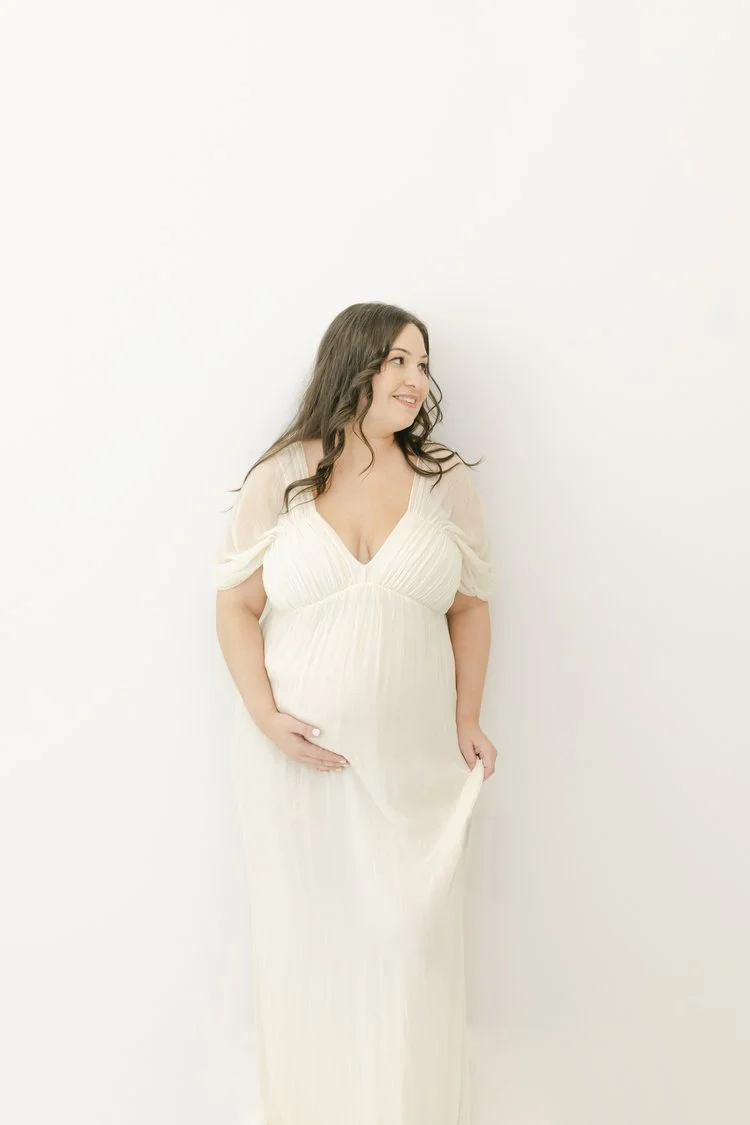 A pregnant woman with long wavy brown hair wearing a flowing white dress, standing against a plain white background, smiling and gently resting one hand on her belly.