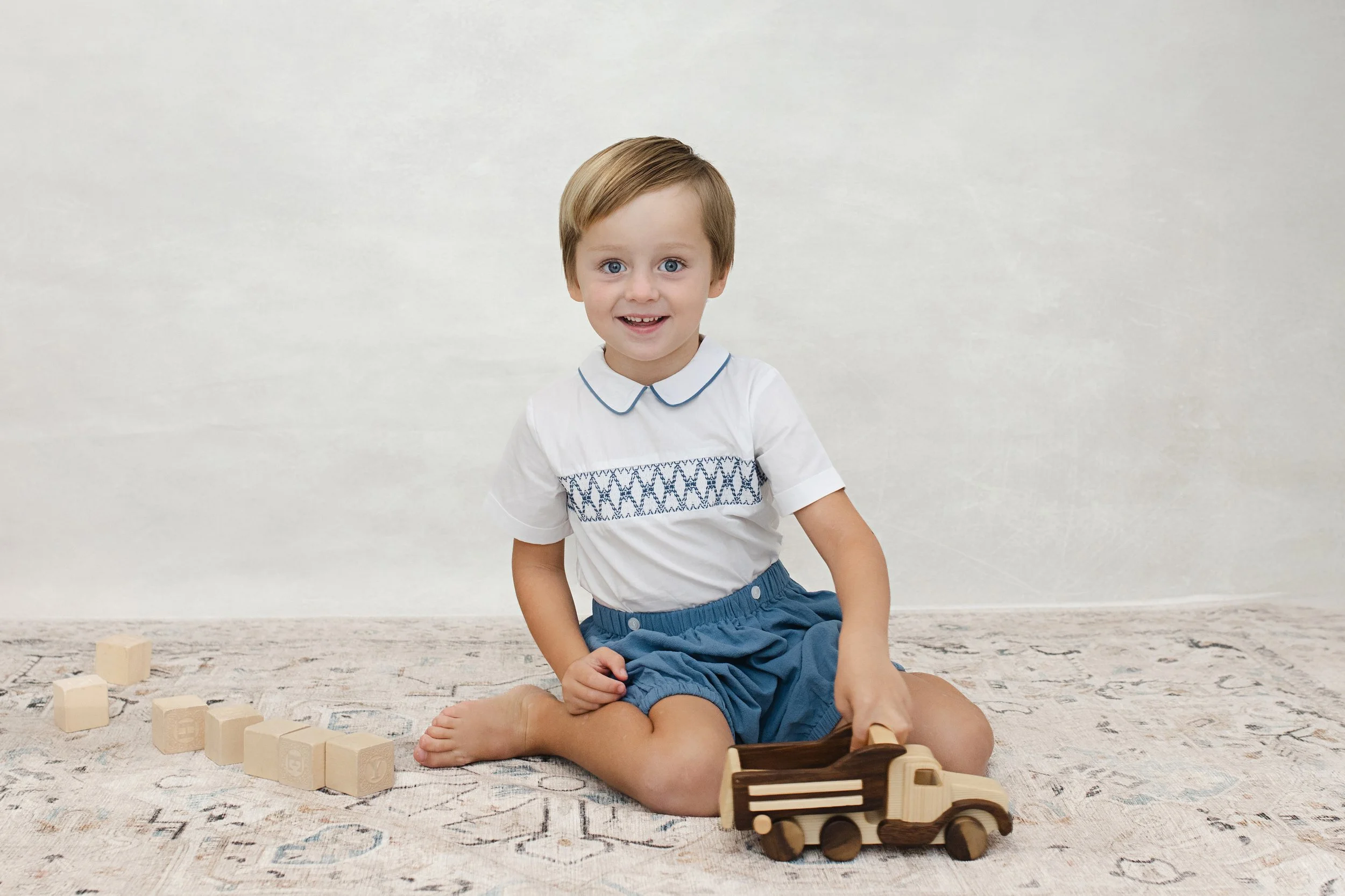 Young boy with light brown hair and blue eyes playing with wooden toy truck and blocks on a patterned rug, smiling at the camera.