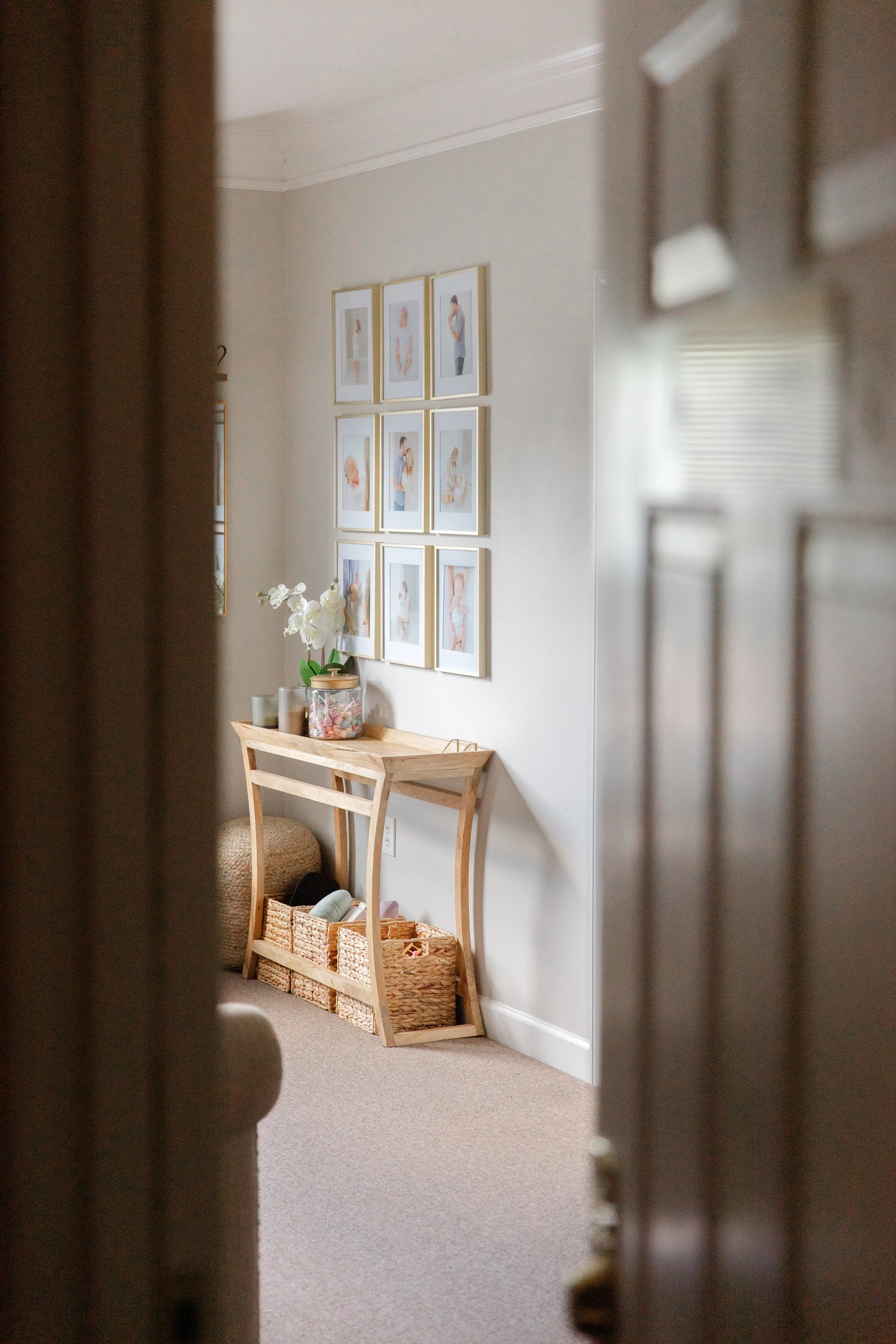 A corner of a room with a light-colored wall decorated with a nine-panel photo gallery. Below the photos is a small, light wood table with a potted orchid and some candles on it. Basket storage is seen underneath, containing various items.