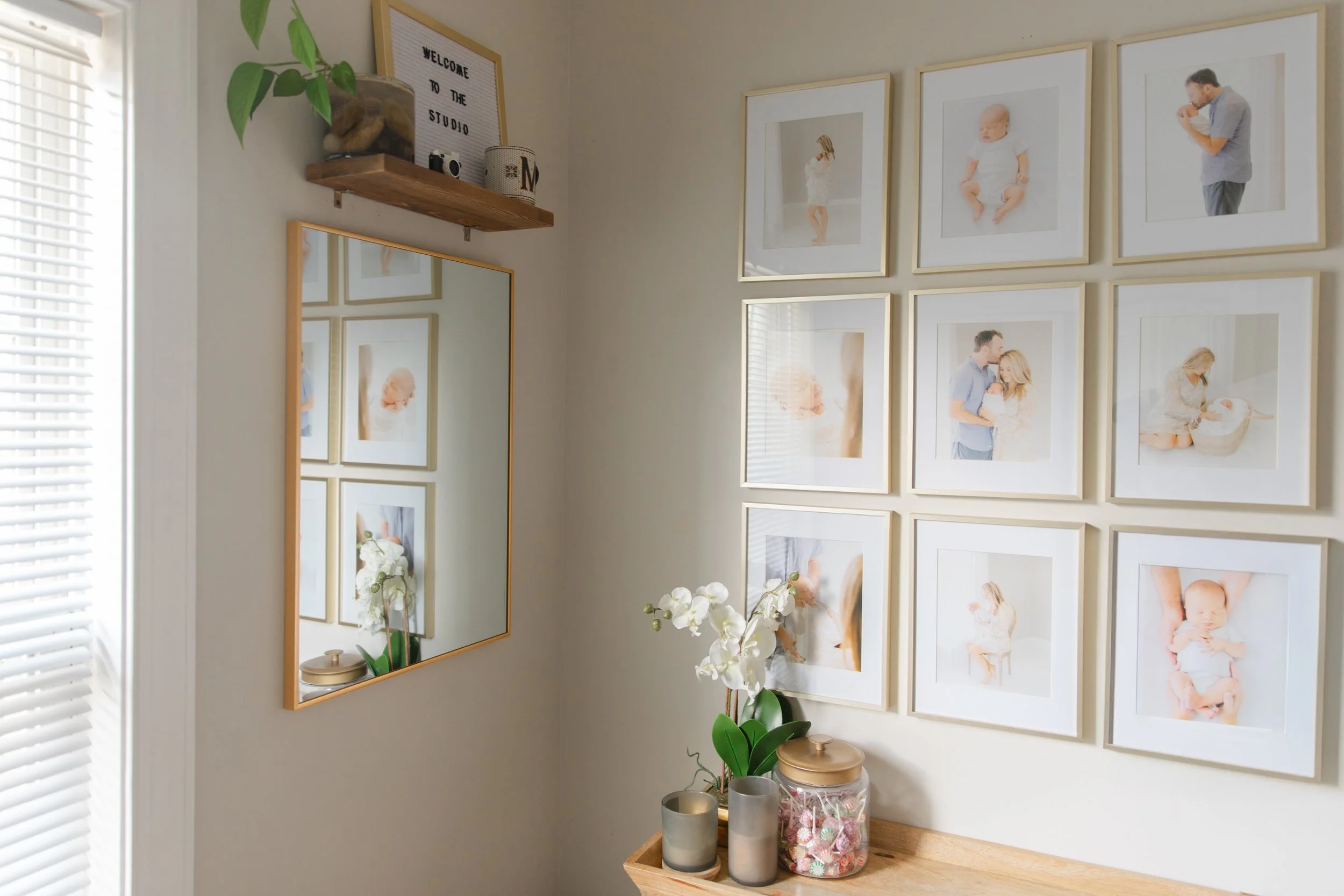 A wall decorated with a collage of family photos, a mirror, a small shelf with plants and decorative items, and a table with plants, candles, and a jar of colorful candies.