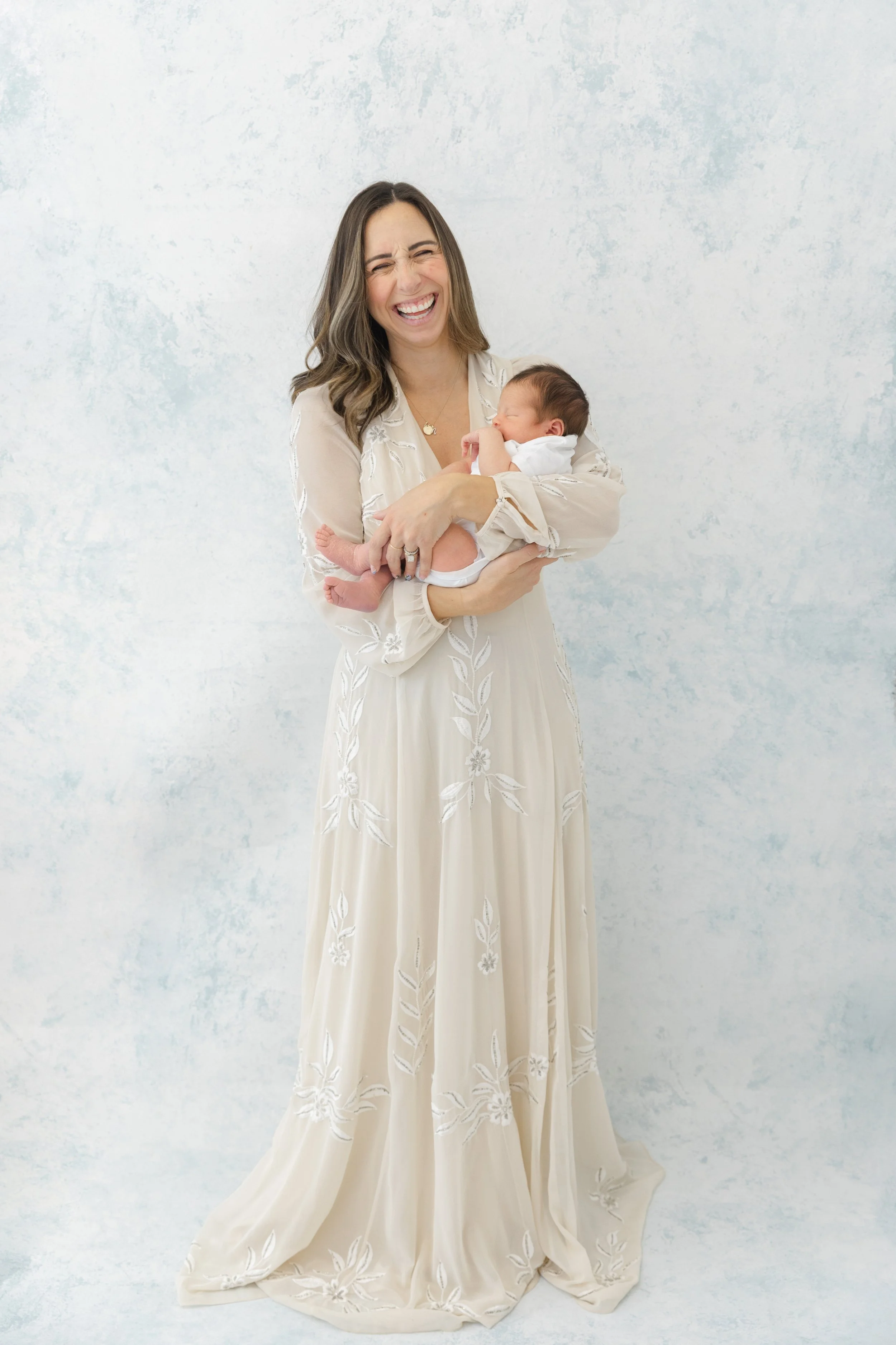A woman in a cream-colored, embroidered dress holds a sleeping baby in her arms, smiling with her eyes closed.