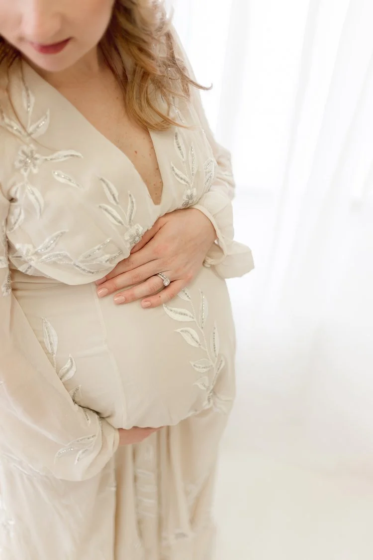 A pregnant woman wearing a cream-colored dress with embroidered leaves, holding her belly with one hand and resting her other hand on top, in front of a light-colored curtain background.