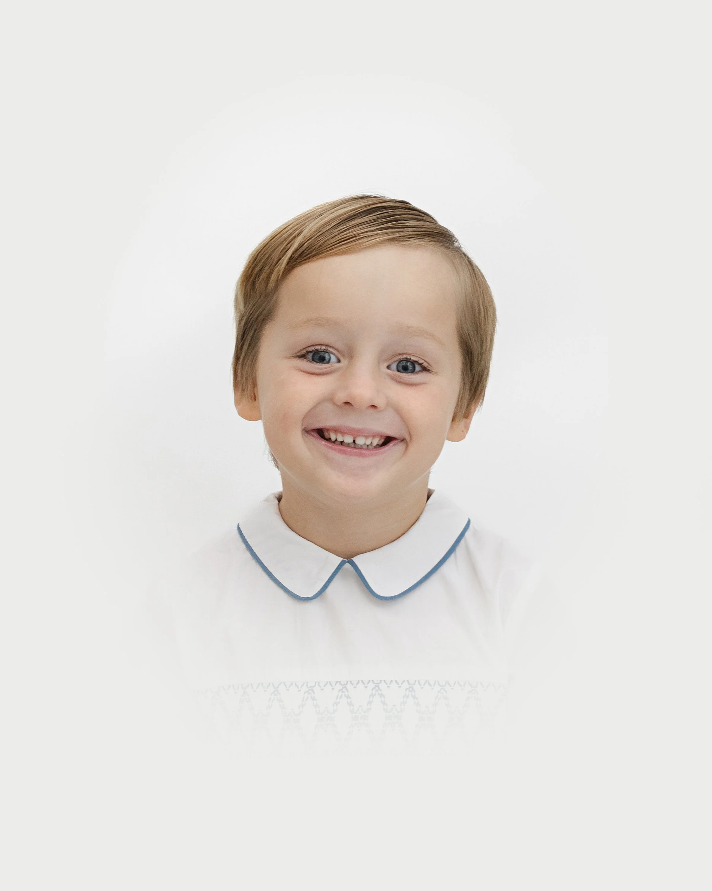 A young boy with light brown hair and blue eyes smiling, wearing a white shirt with a collar and a blue trim, against a white background.