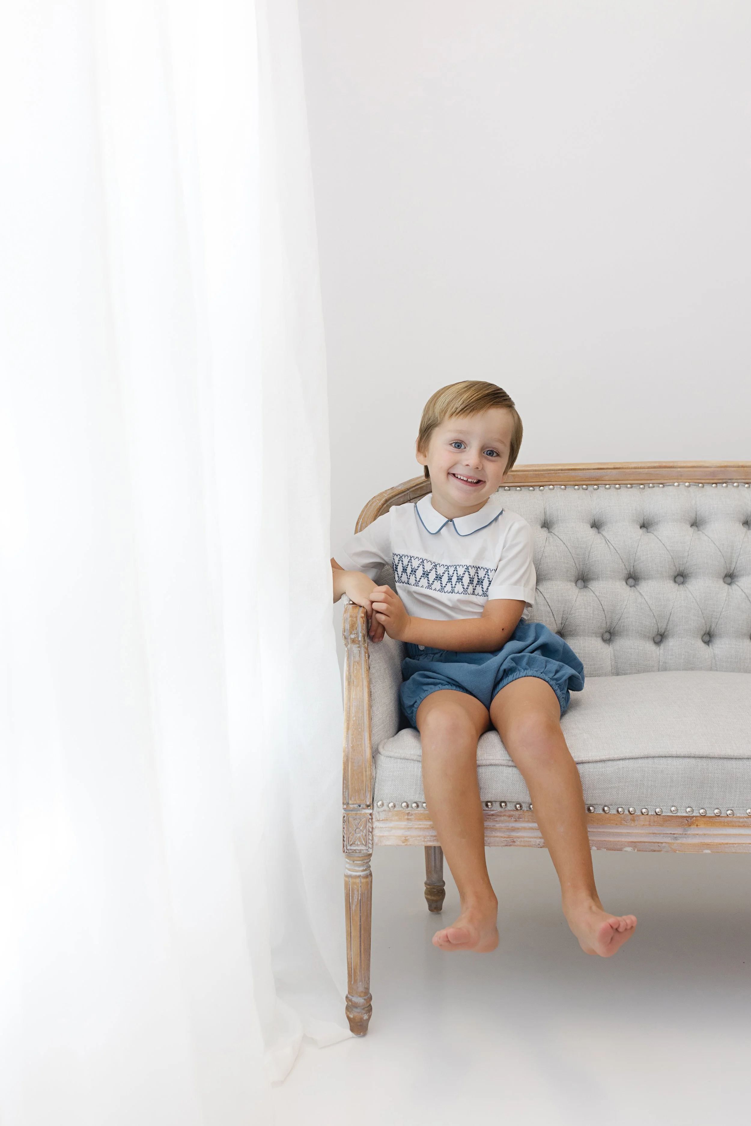 A young boy with light brown hair sitting on a vintage-style beige sofa with a tufted back, smiling and looking at the camera.