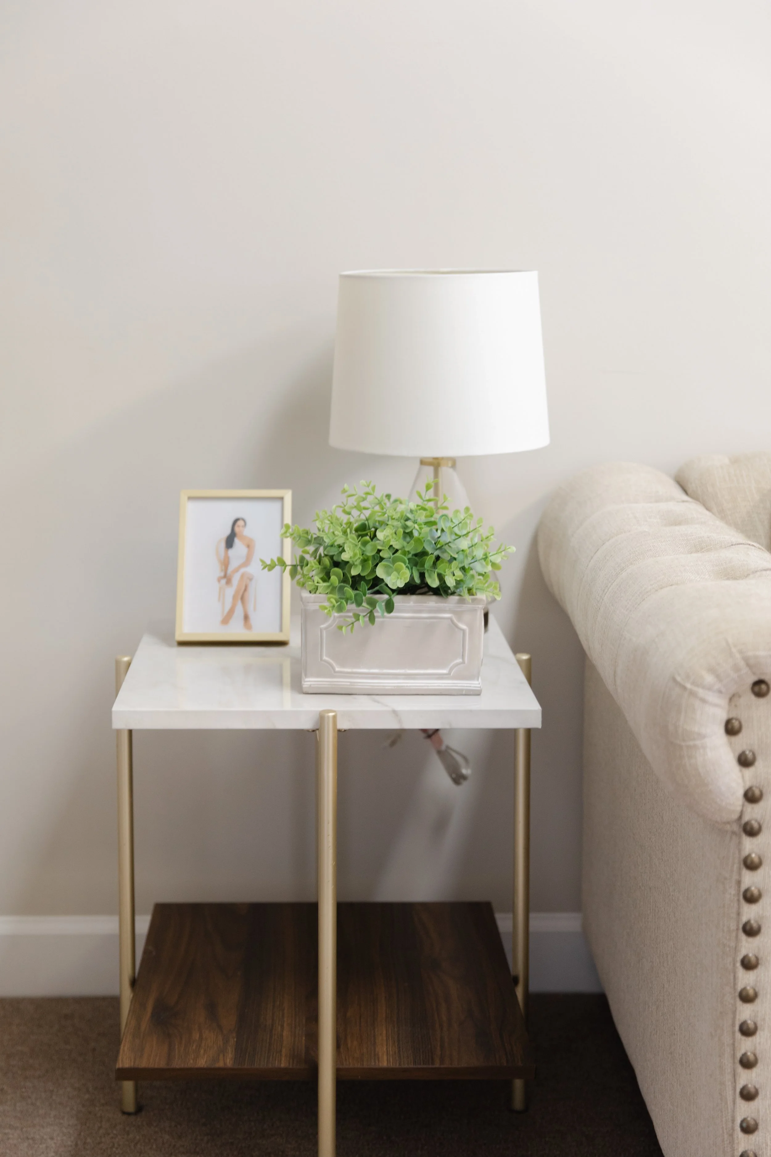 A beige upholstered sofa with decorative nailhead trim next to a side table with a white marble top, gold legs, and a dark wooden bottom shelf. The table holds a white lampshade, a framed photograph of a woman in white, and a rectangular silver planter with green artificial plants.