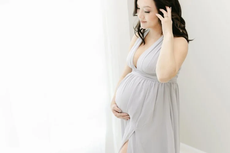 Pregnant woman in a light gray dress standing near a white wall, gently touching her belly and smiling softly.