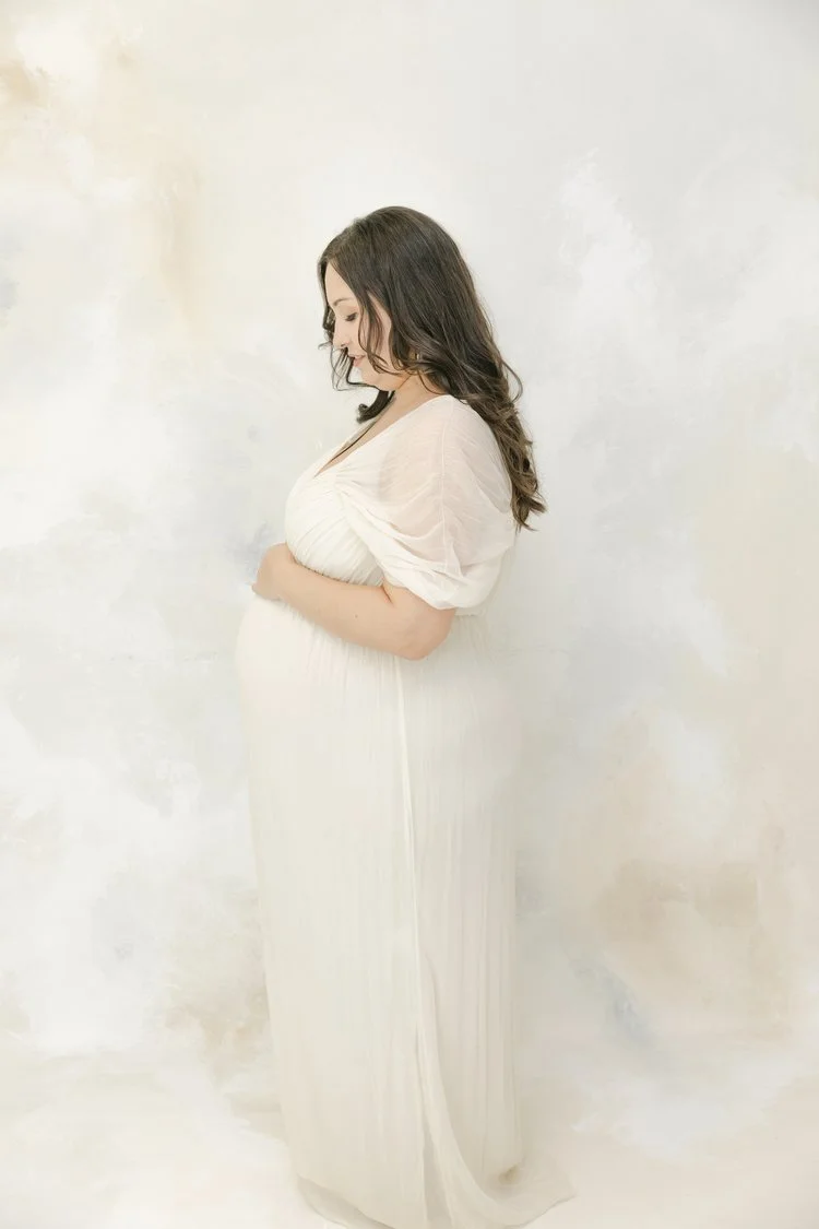 A pregnant woman with long dark hair in a white dress standing sideways against a plain wall, looking down, smiling gently.
