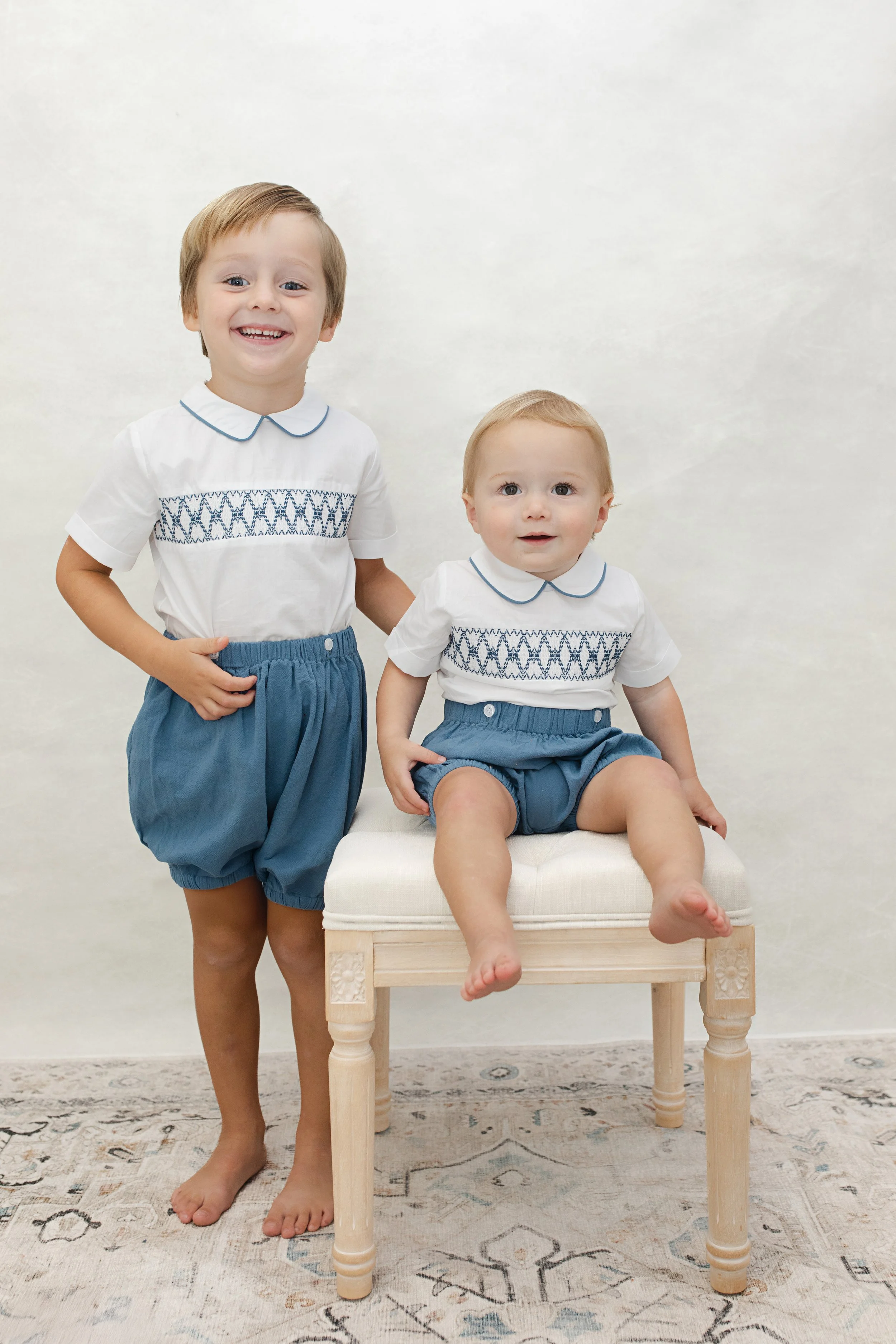 Two young boys in matching white shirts with blue embroidery, blue shorts, one standing and one sitting on a small upholstered bench, smiling and posing for a photo.