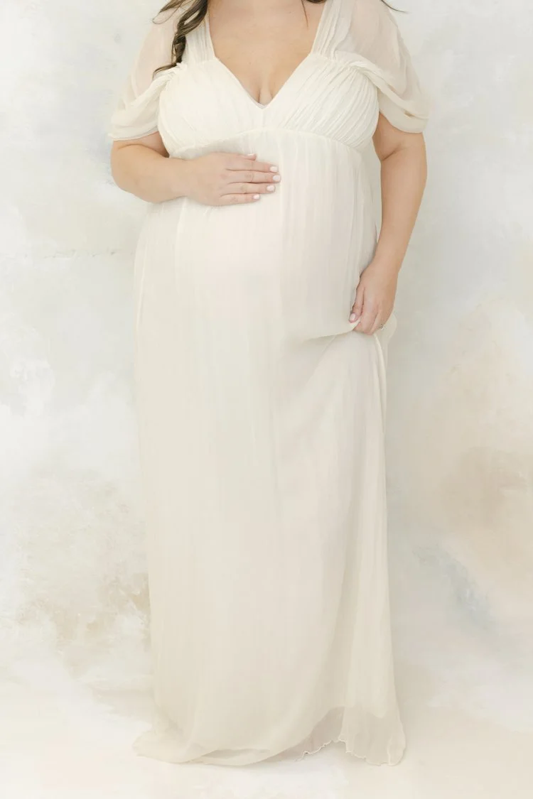 A woman wearing an ivory-colored, flowing maxi dress with pleated details on the bodice, standing against a light-colored wall. She is placing one hand on her pregnant belly.