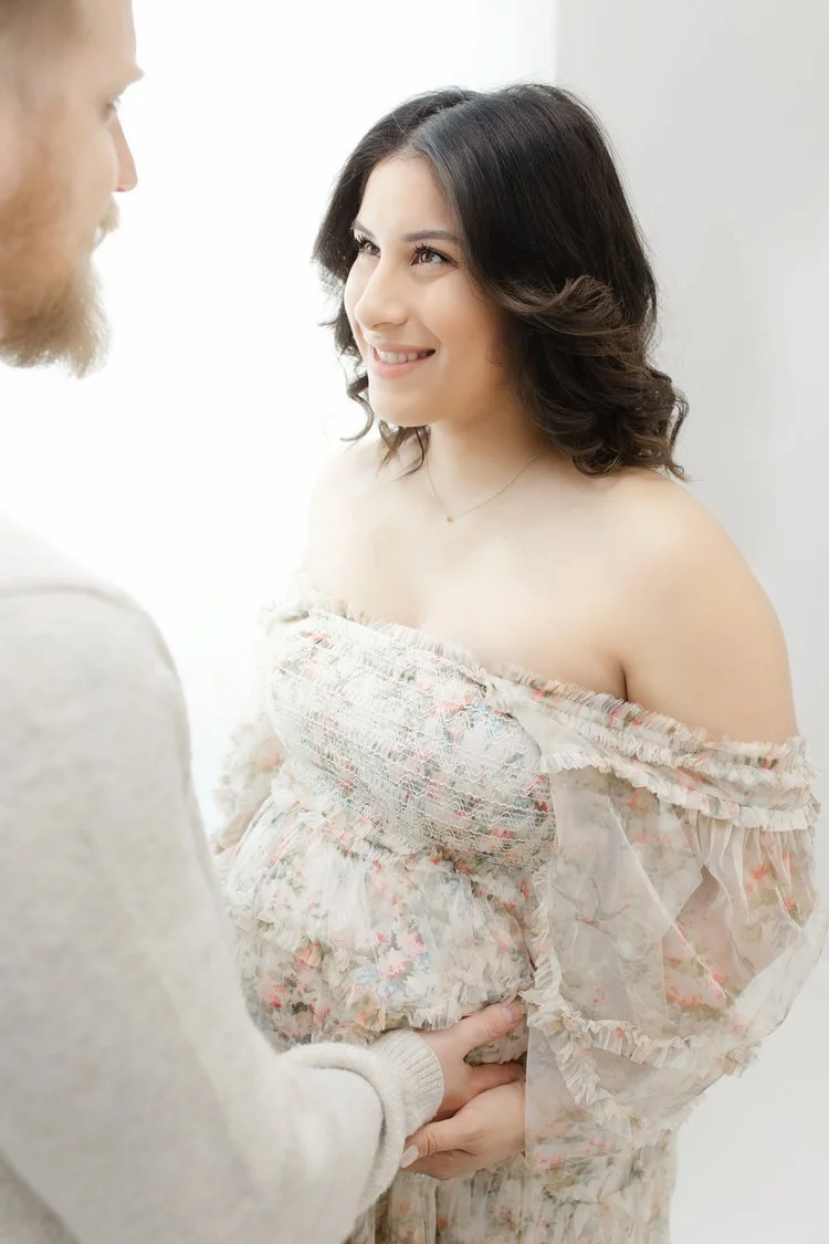 A pregnant woman with dark, wavy hair smiling at a man with a beard, in a light-colored room. The woman is wearing an off-the-shoulder floral dress, and the man is holding her pregnant belly gently.