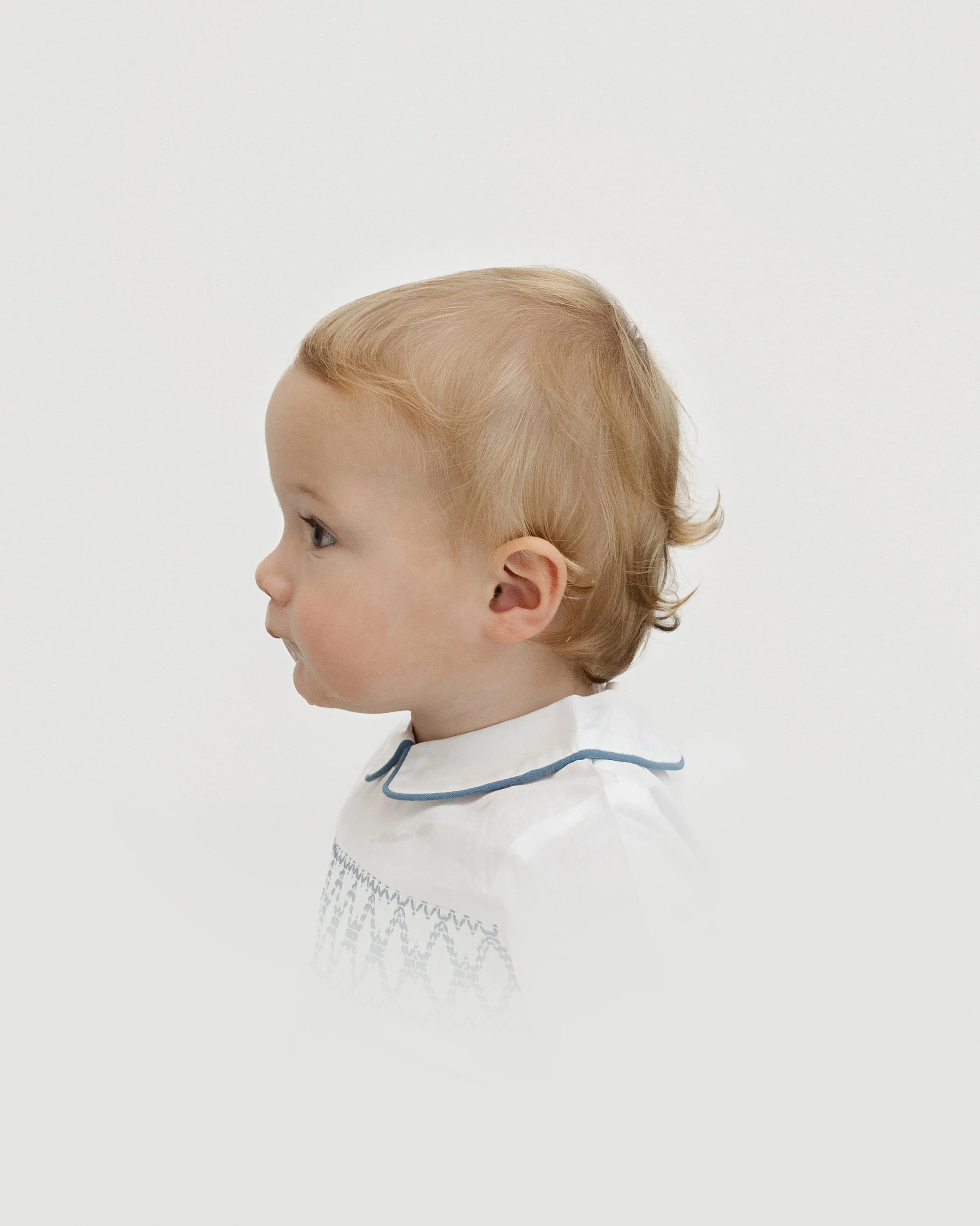 Side profile of a young blonde child wearing a white shirt with a blue trim on the collar, against a plain white background.