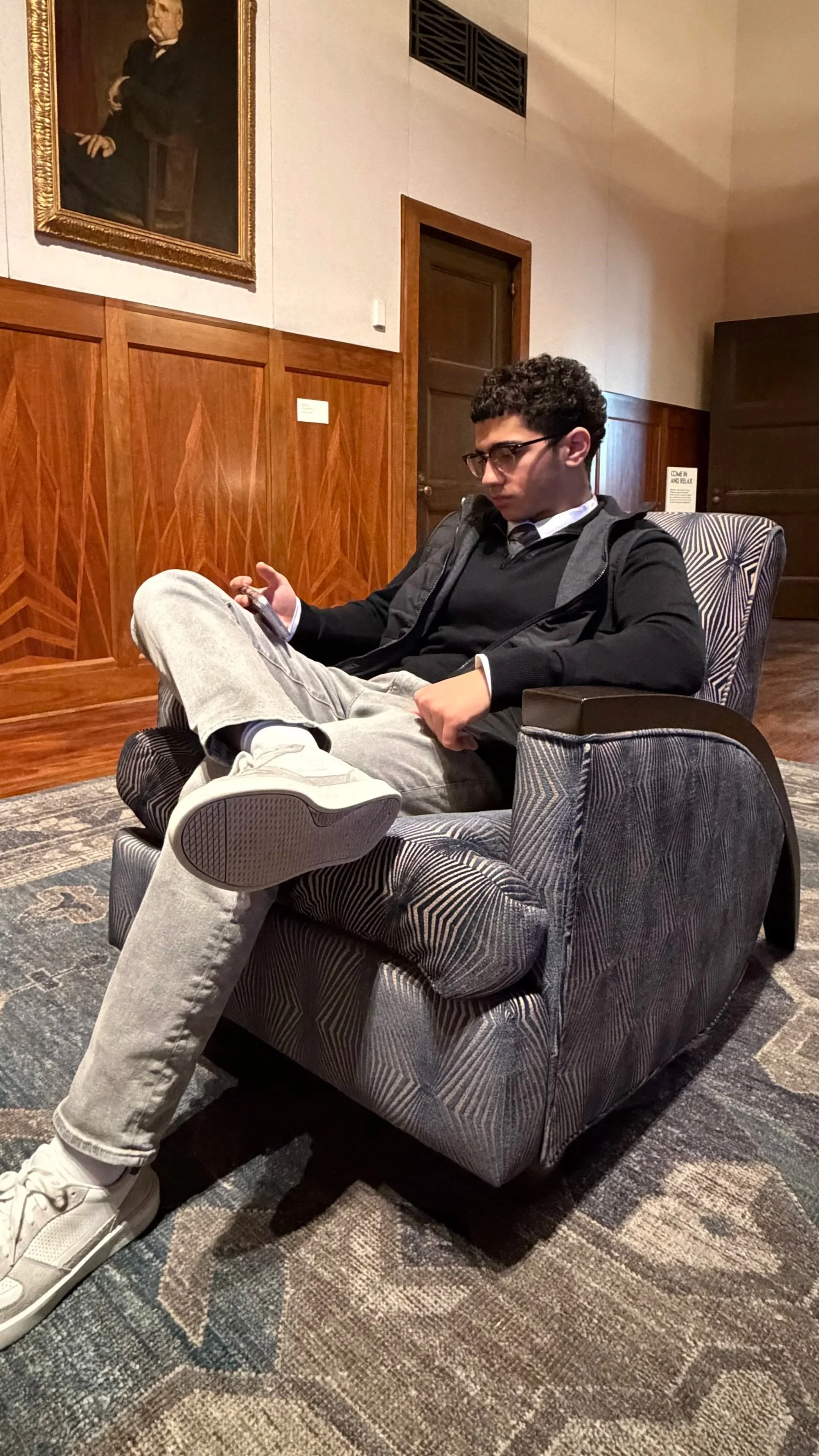 A young man with glasses, wearing a black jacket and gray pants, is sitting in a patterned armchair in a room with wooden paneling. He is looking at his phone with one leg crossed over the other. Behind him, there is a wooden door and an art portrait on the wall.