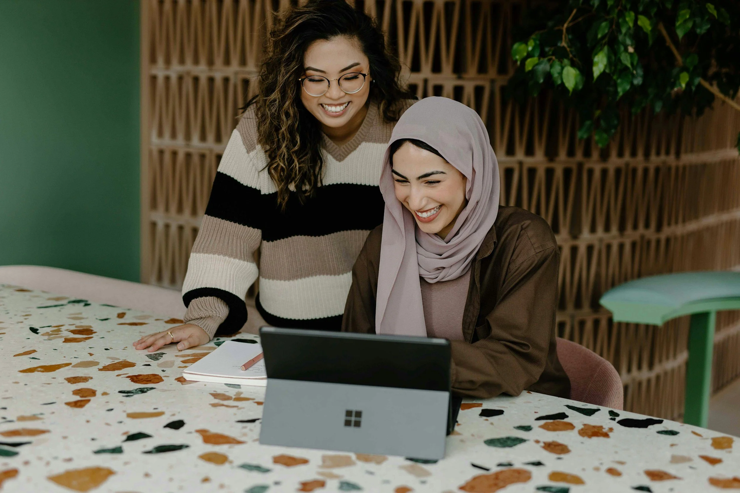 Two women smiling and looking at a tablet together at a table in an indoor setting.