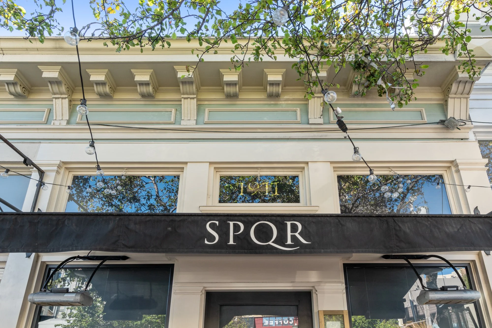 The exterior of a building with a sign that reads 'SPQR' and decorative trim above the windows. String lights are hung across the space, and the building features three large windows, with reflections of trees and the sky visible in them.