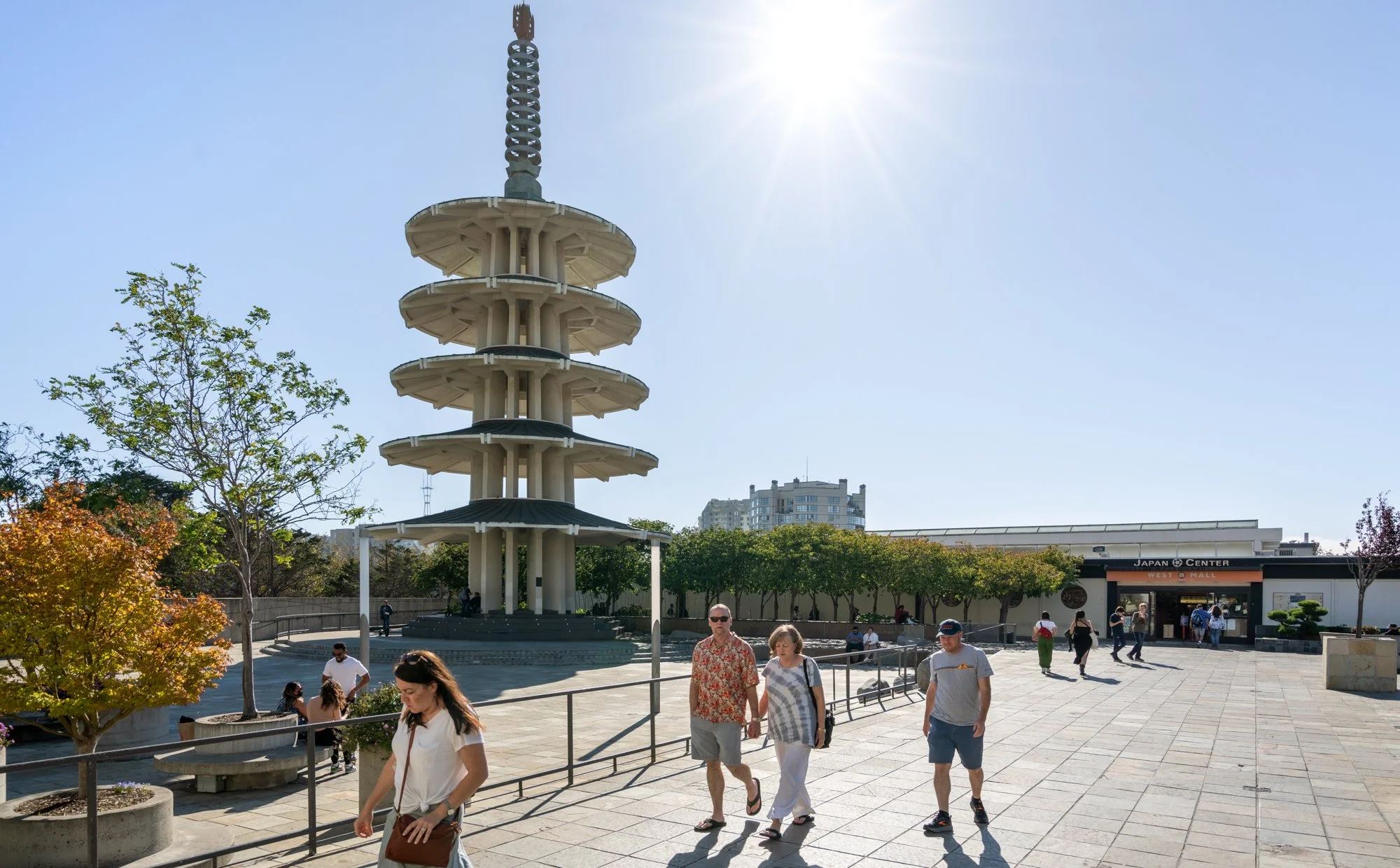 A group of people walking outside in front of a large, multi-tiered viewing tower with trees around it and a building labeled Japan Center in the background.