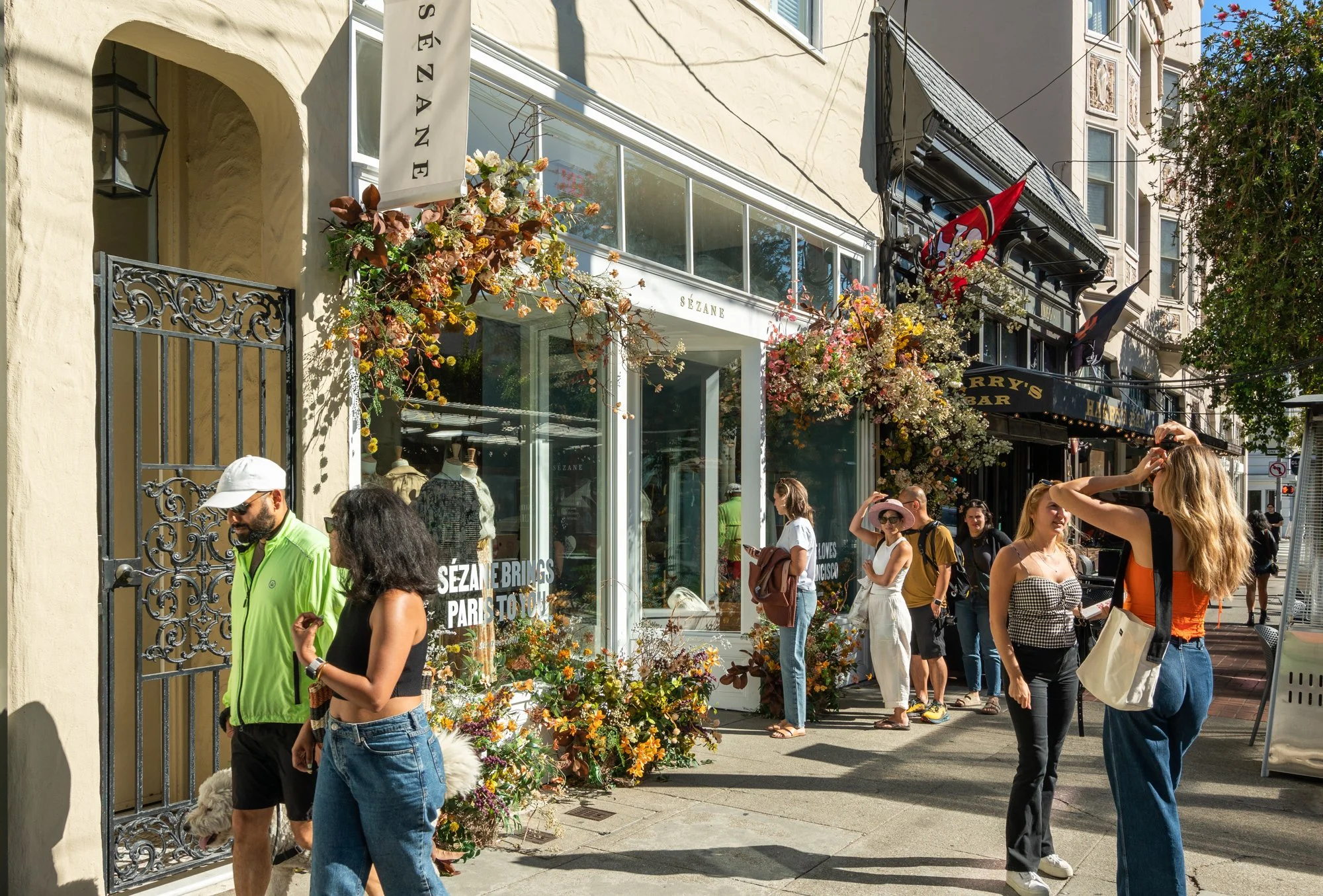 People standing outside a flower-decorated storefront on a sunny day, some looking at their phones and others chatting.