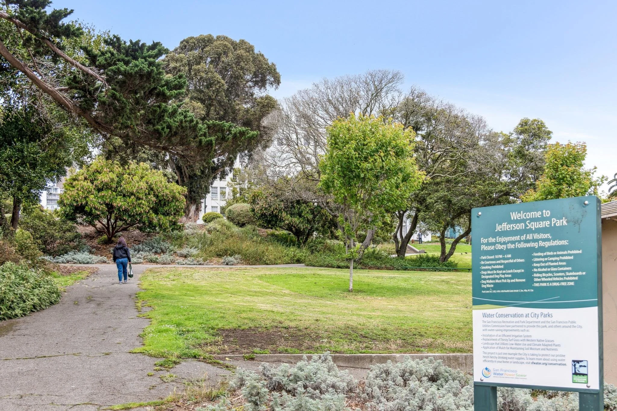 A person walking along a paved path in Jefferson Square Park with trees and shrubs, a welcome sign, and buildings in the background.
