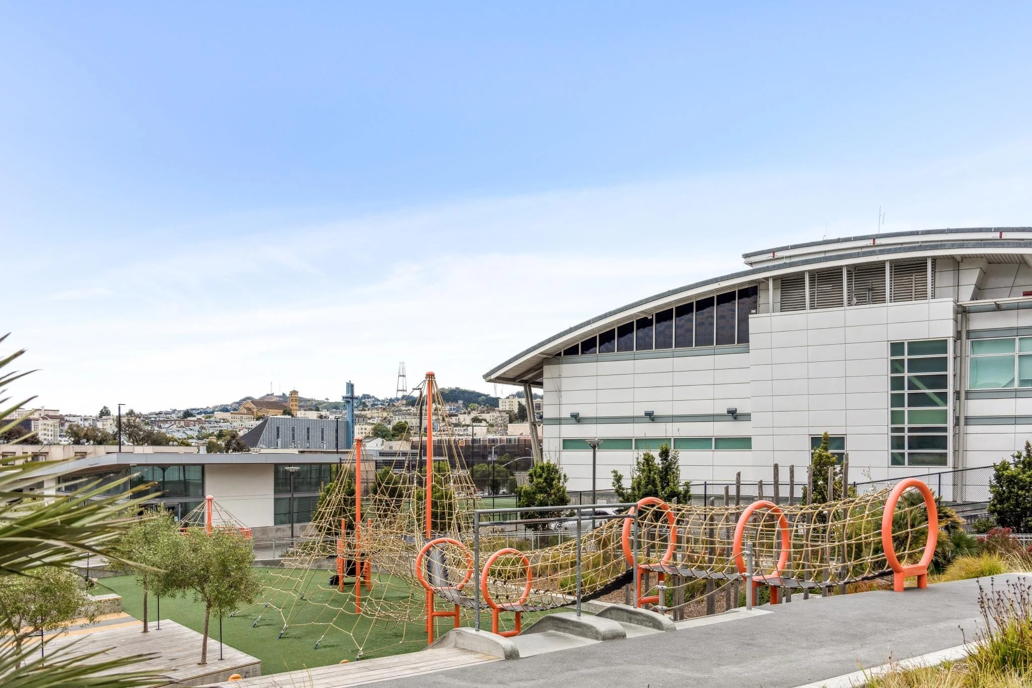 Modern urban playground with climbing structures and a building in the background, situated in a landscaped area with trees and plants.