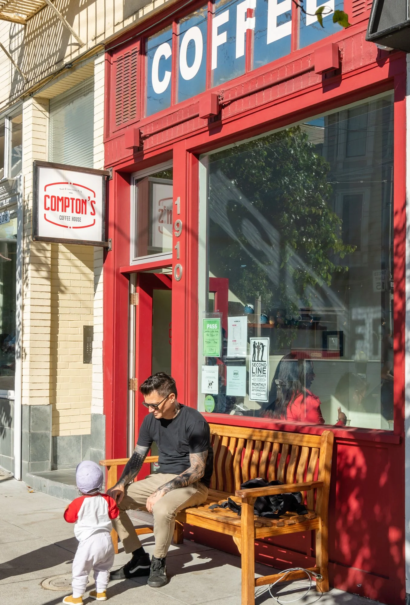 A man sitting on a wooden bench outside Compton's Coffee House, interacting with a small child in a white outfit and hat, on a sunny day.