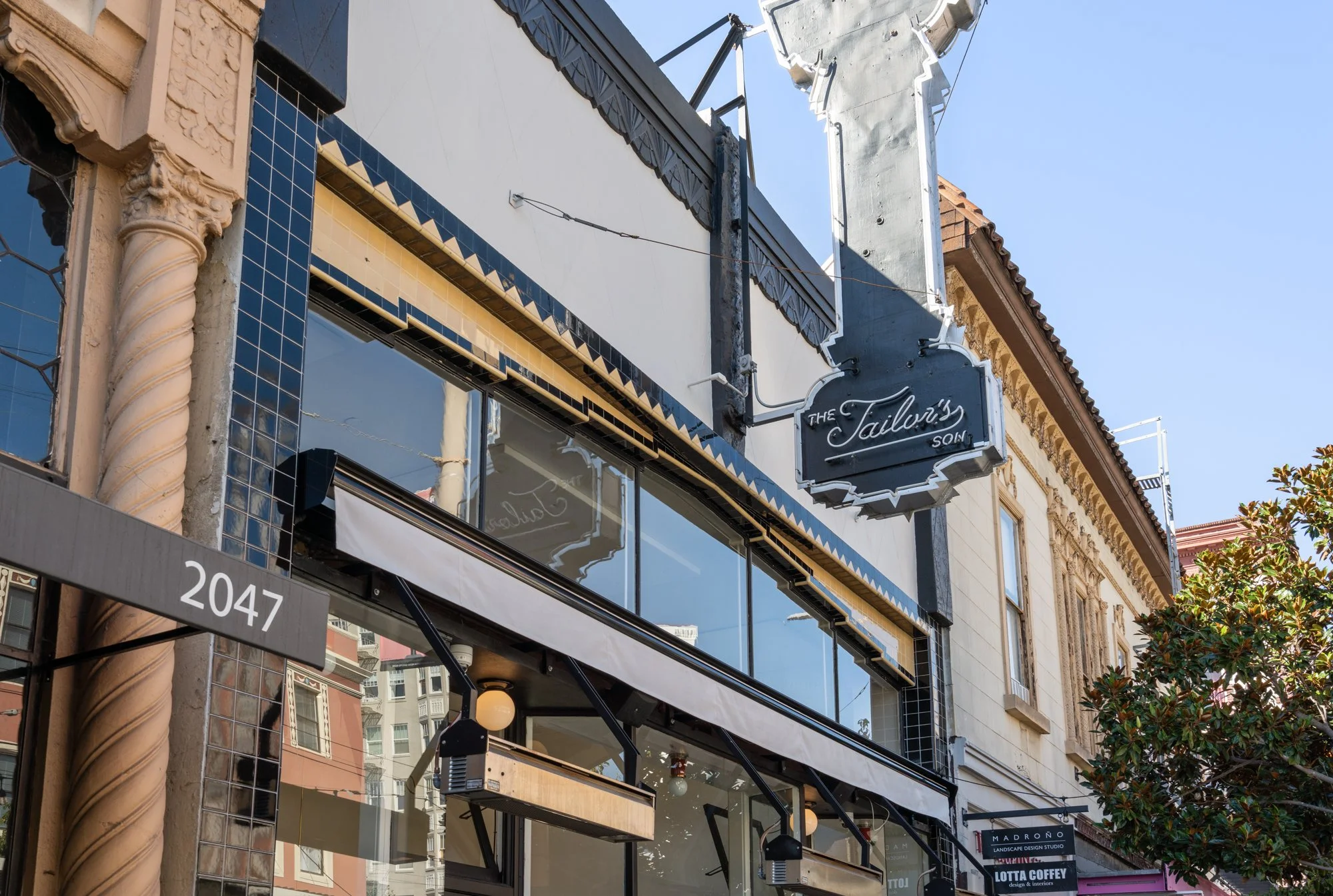 Close-up of a building facade with a sign reading 'The Tailor's Son,' large windows, and decorative architectural details on a city street.