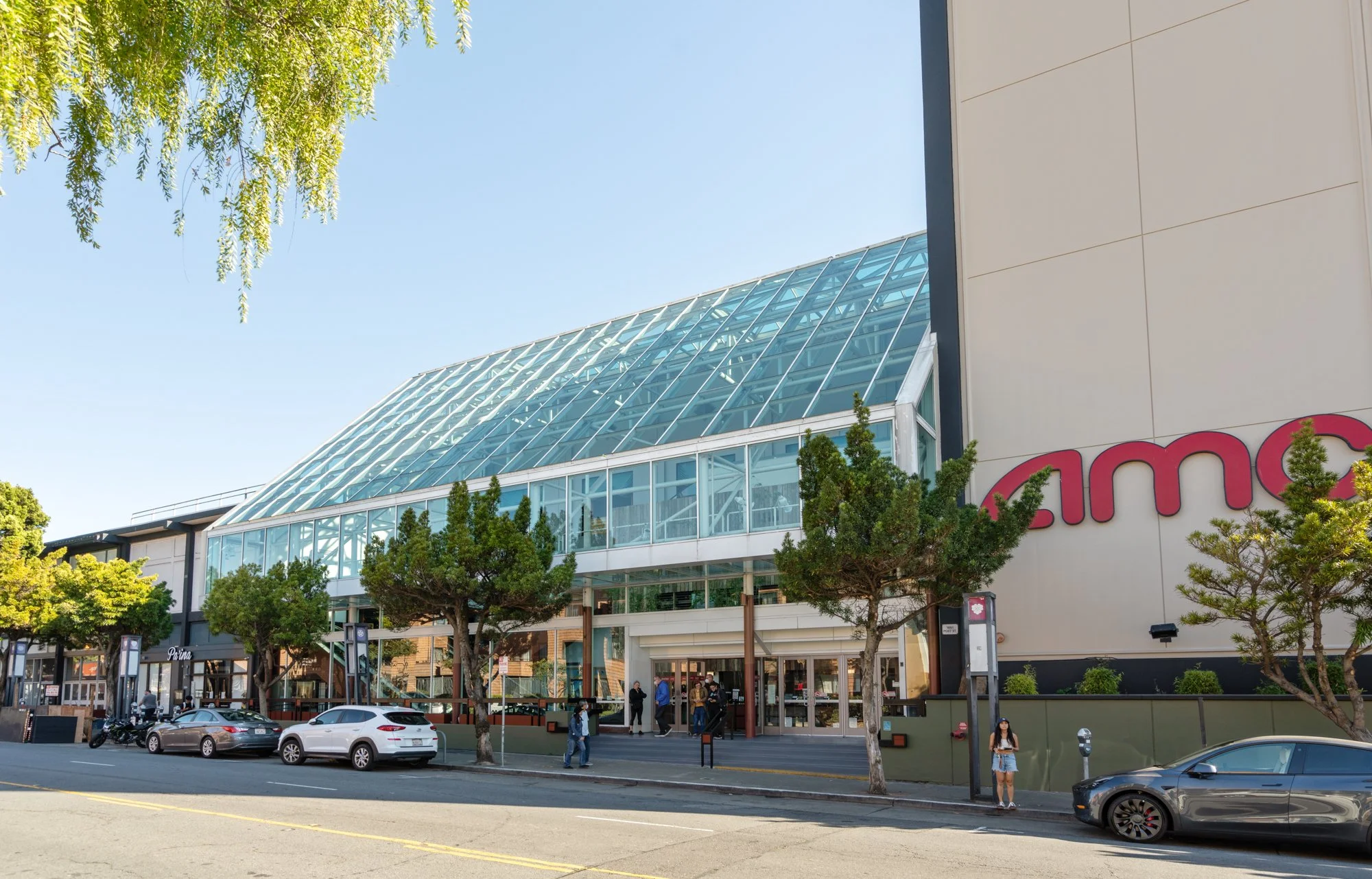 Exterior of a modern shopping mall with glass entrance and trees lining the sidewalk, several parked cars, and people walking outside.