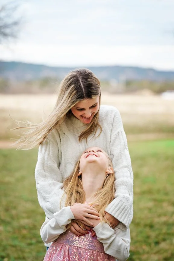 Big sister in a cream cable knit sweater hugging her laughing little sister from behind in an open field, showing texture in family photo outfits.