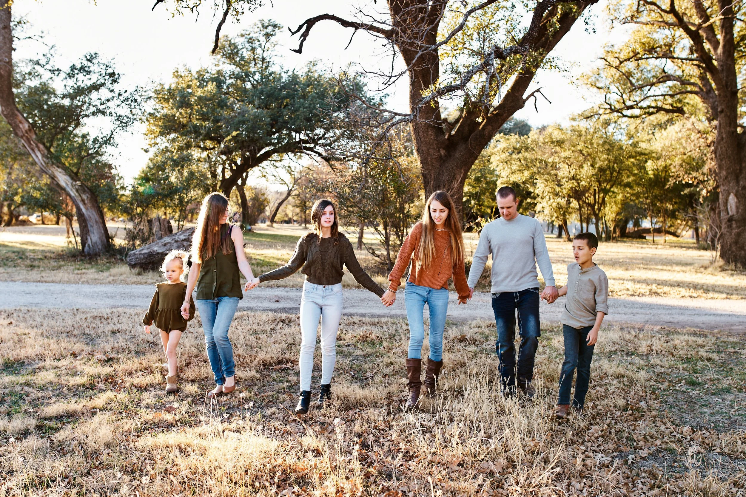 Family of six holding hands and walking under oak trees in fall tones of rust, olive, and denim during an outdoor session near Abilene, TX.
