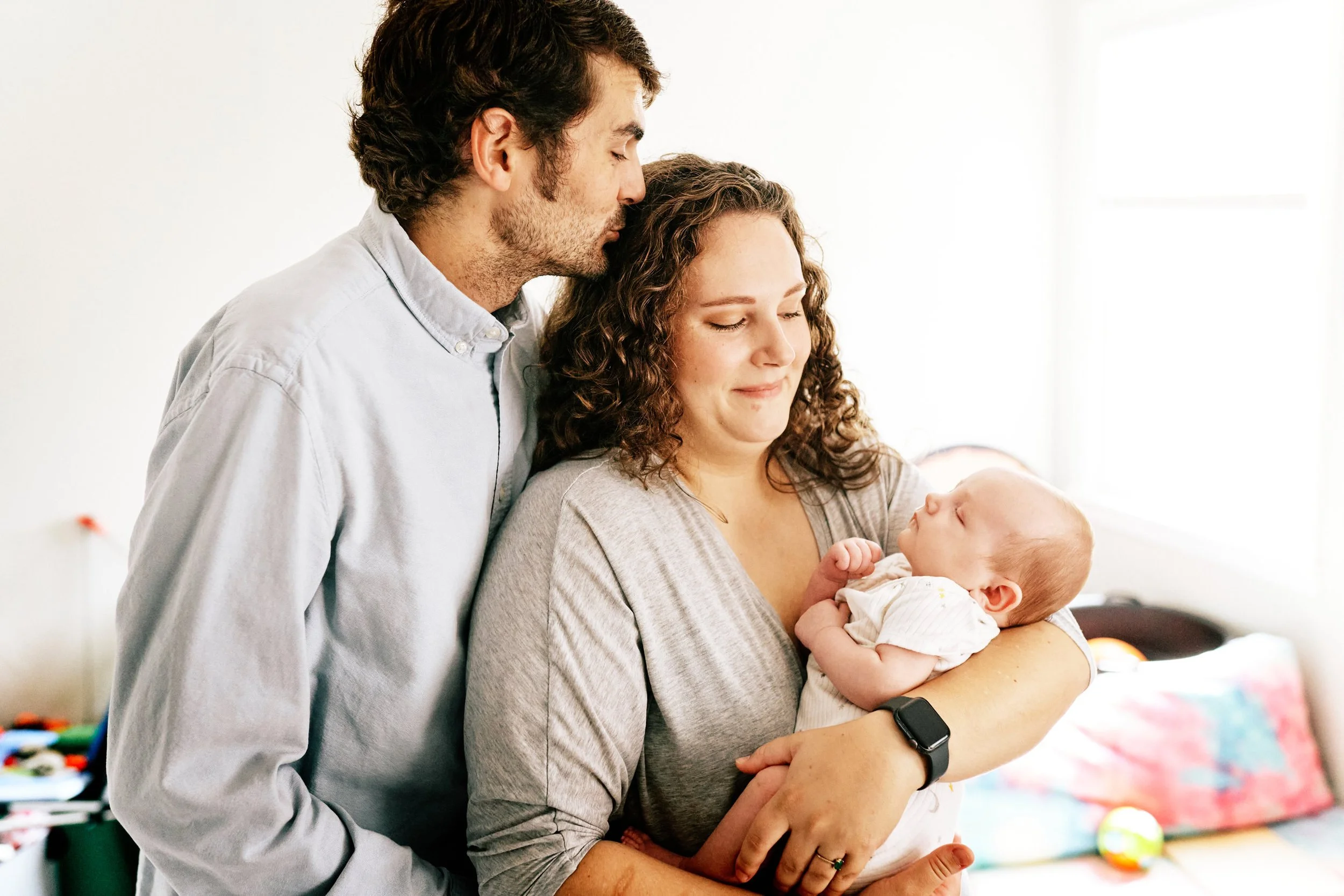 New mom cradling newborn in a brightly lit room with a window. Husband is holding mom from behind and kissing her on the head.