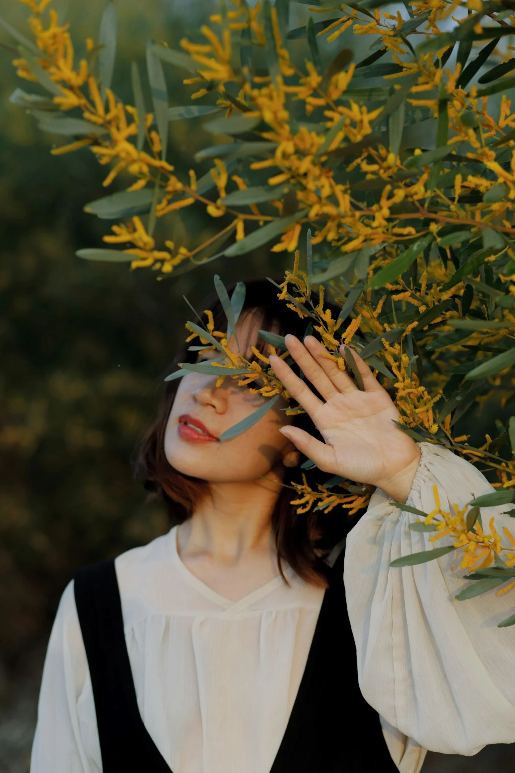 Eine junge Frau mit dunklen Haaren und rotem Lippenstift, die eine weiße Bluse trägt, steht im Freien vor einem Busch mit gelben Blumen und grünen Blättern, während sie eine Hand hebt, um das Blatt vor ihrem Gesicht zu halten.