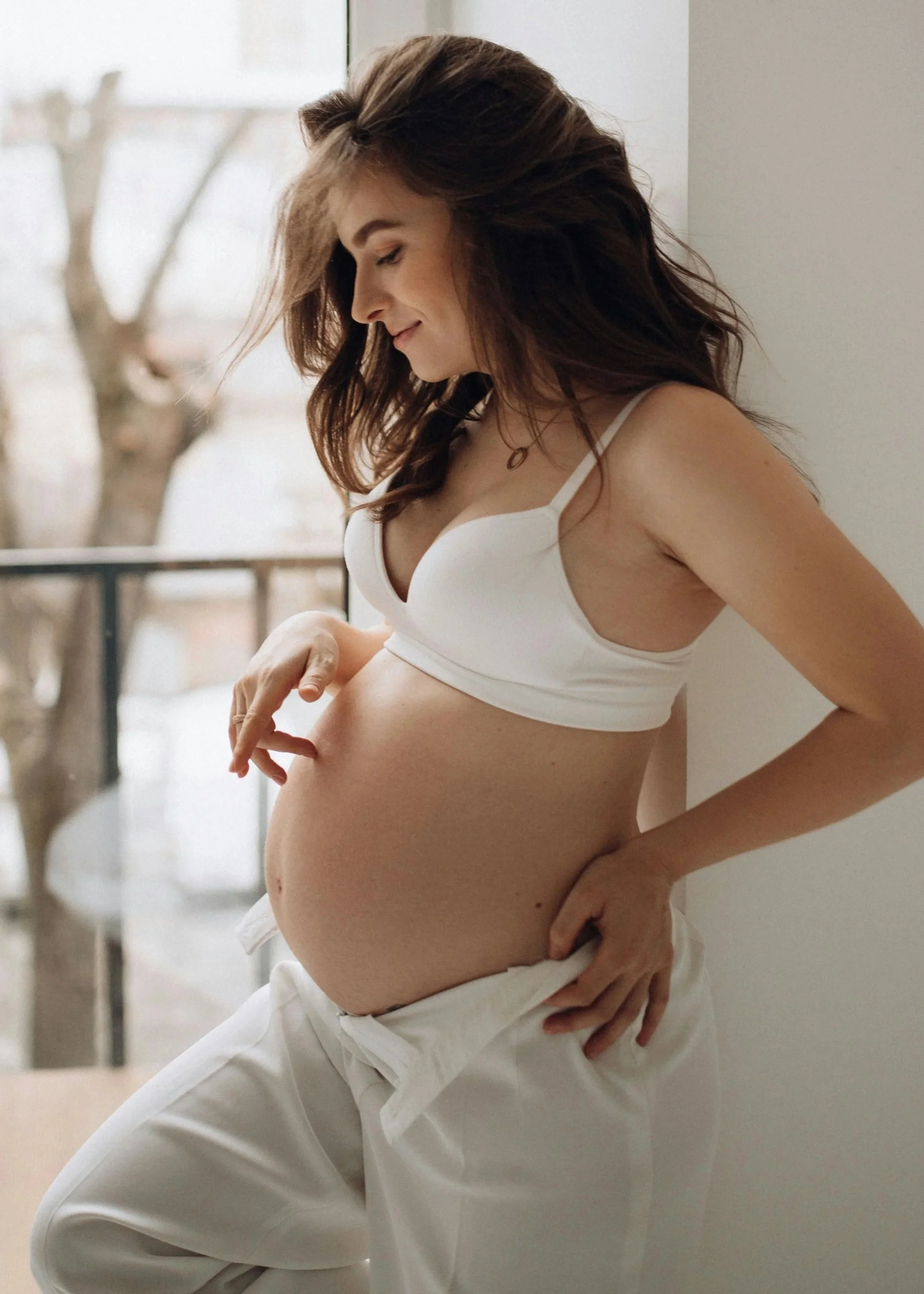 Schwangere Frau in weißem Hemd und Hose steht vor Fenster, lächelt, während sie ihren Bauch berührt.
