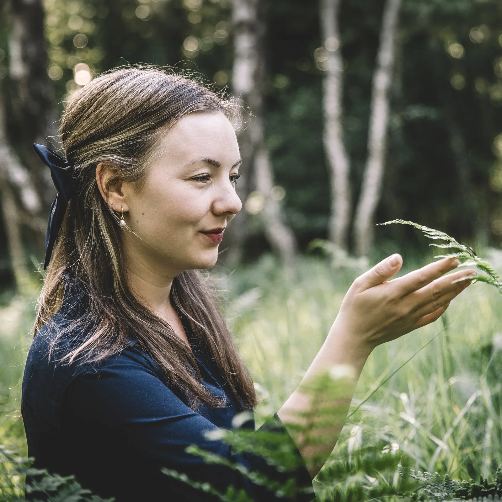 Eine junge Frau mit braunen Haaren, die auf einer Wiese in einem Wald steht und eine Pflanze betrachtet.