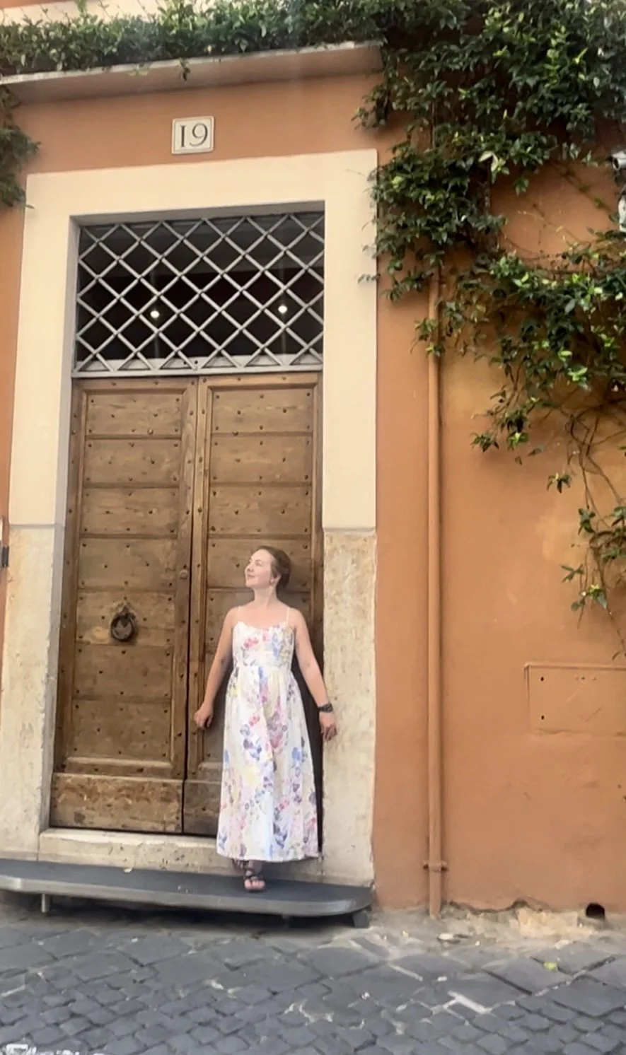 Junge Frau in einem weißen, bunten Kleid steht vor einem großen Holztor an einer orangefarbenen Wand, umgeben von grünen Pflanzen.