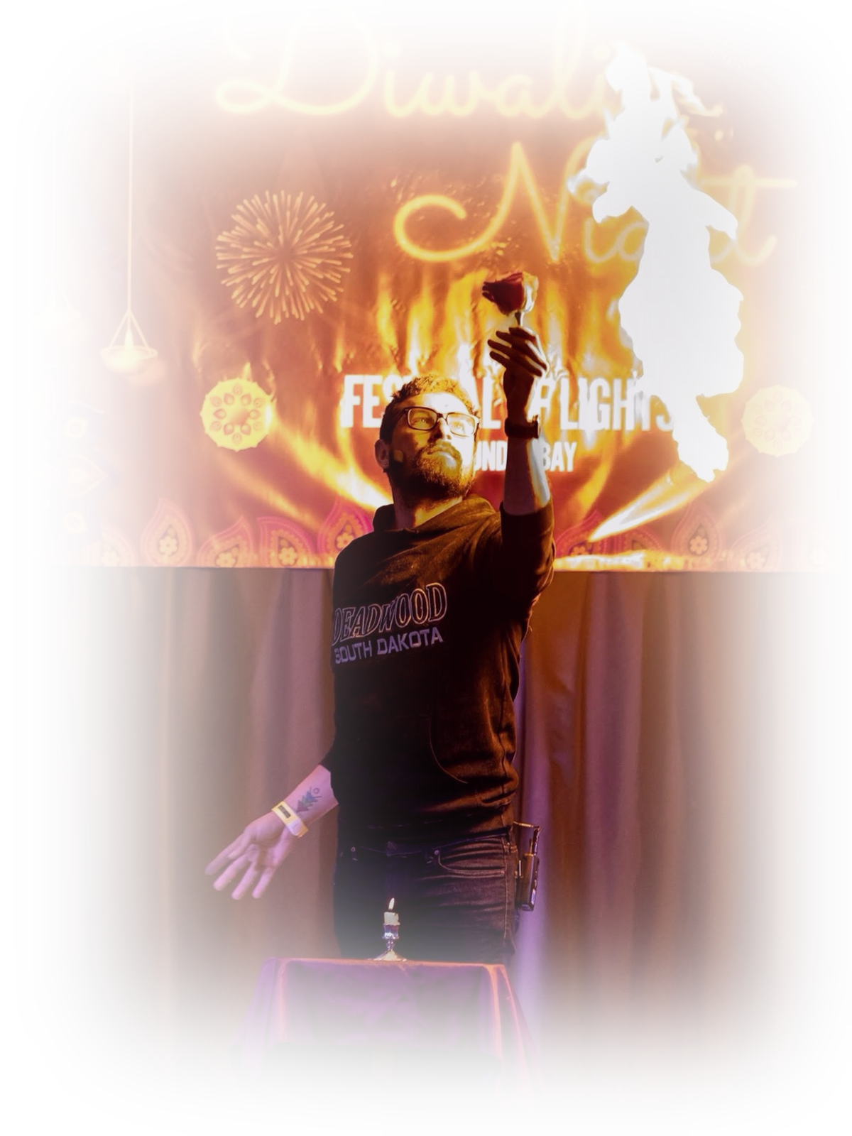 A man wearing glasses and a South Dakota-themed hoodie holding a rose in front of a Diwali and Christmas event backdrop with fireworks, lanterns, and decorative patterns. There is a small table with a lit candle in front of him.